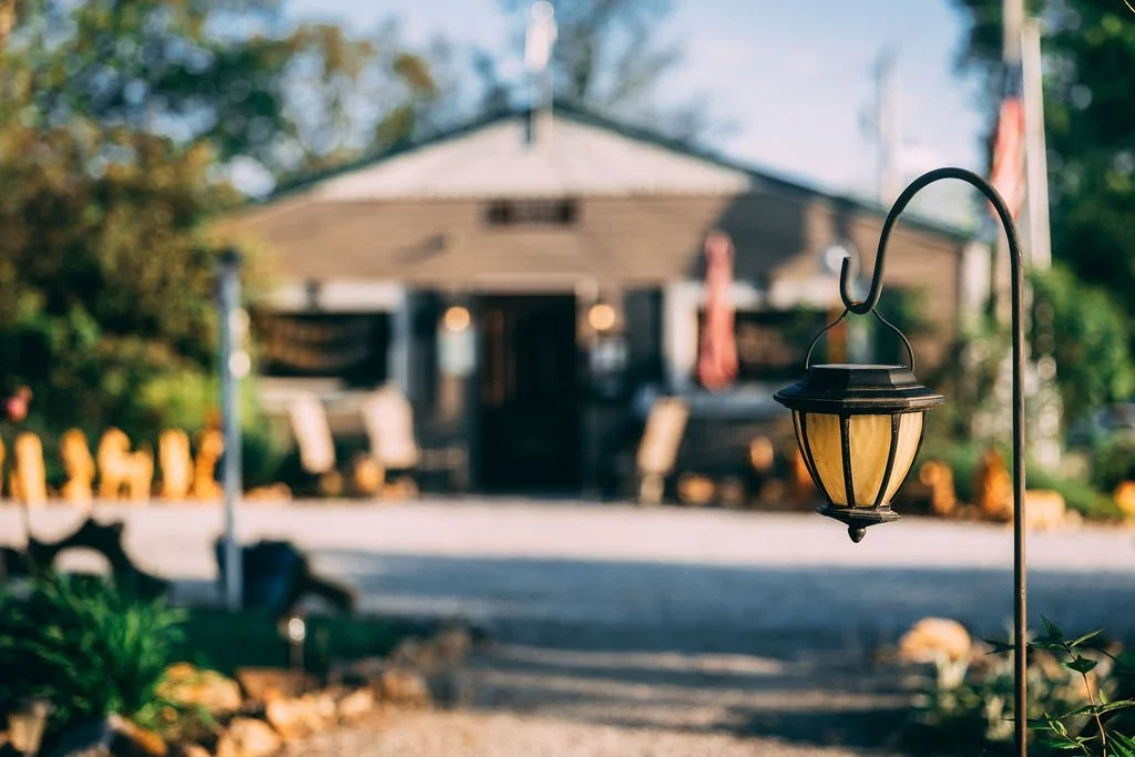 Outdoor scene with a lantern in focus in the foreground and a house with a yard and trees in the background.