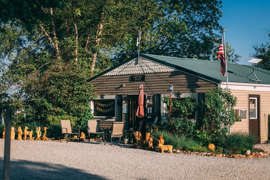 A small, beige building with a green roof labeled 'Lazy Day Office,' surrounded by greenery, outdoor chairs, and garden ornaments, including animal-shaped sculptures.