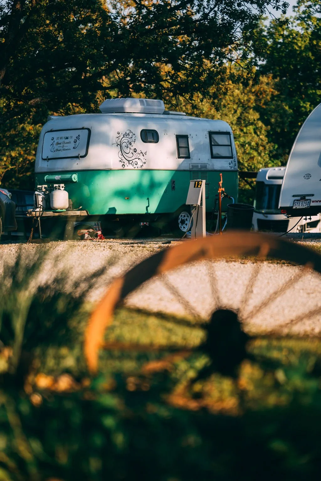A vintage camper trailer with a green lower half and white upper half, decorated with black floral designs, parked outdoors under trees, with other trailers and vehicles nearby.