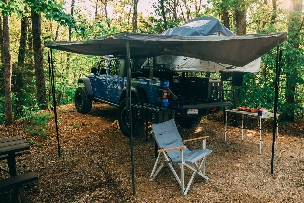 Camping scene in a wooded area with a blue SUV parked under a canopy tent, featuring camping gear and a folding chair.