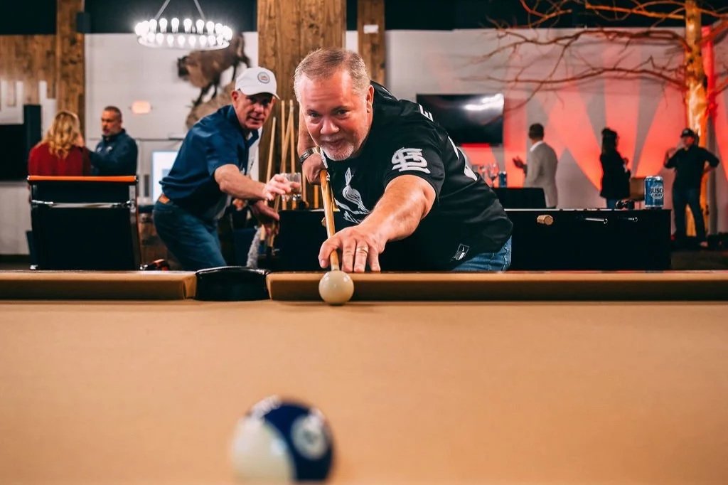 Man in a black sports jersey aiming to hit a cue ball in a game of billiards or pool at a bar or game room, with other people and decorations in the background.
