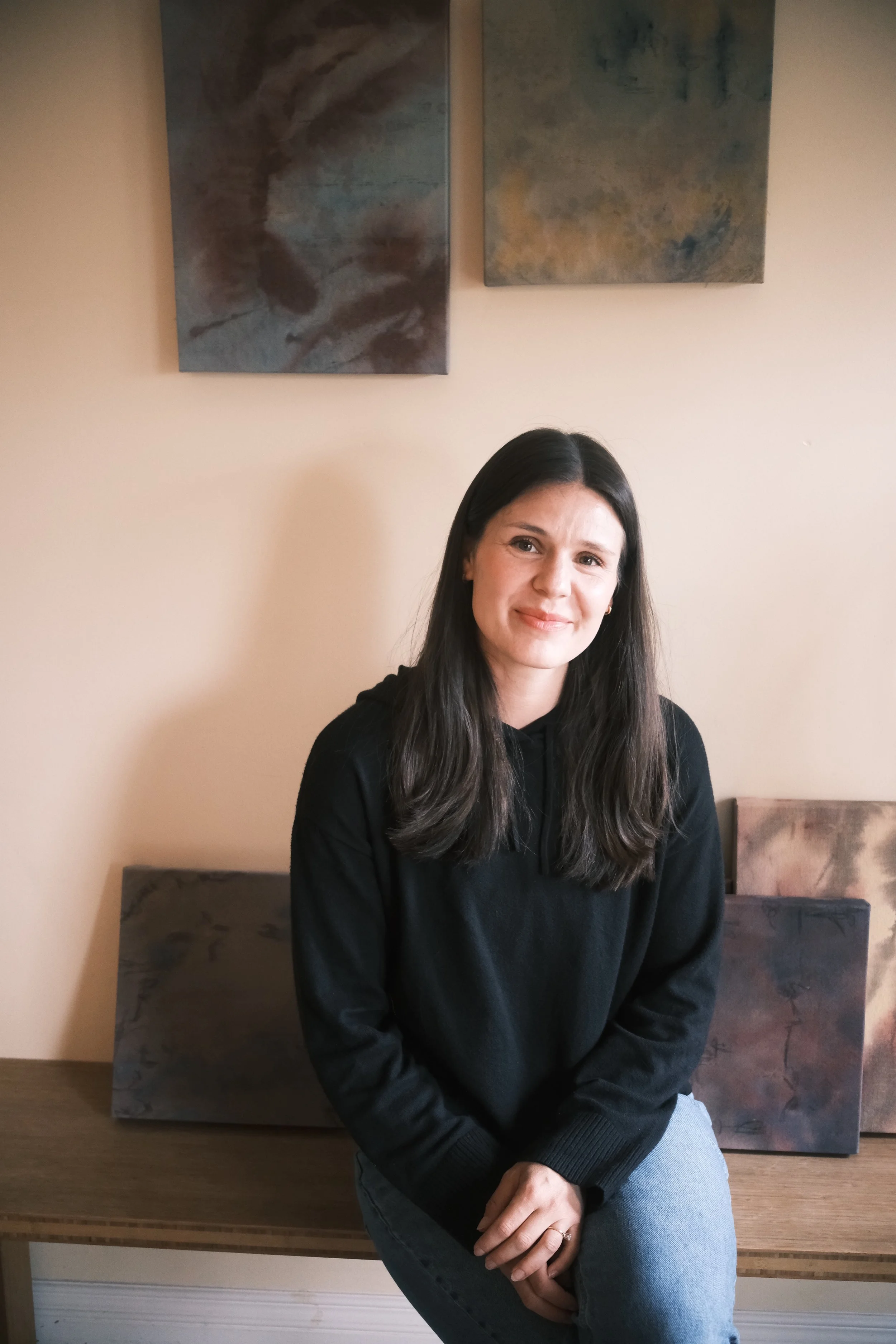 A woman with long dark hair wearing a black hoodie and light blue jeans sitting on a wooden bench in front of a beige wall with two abstract paintings.
