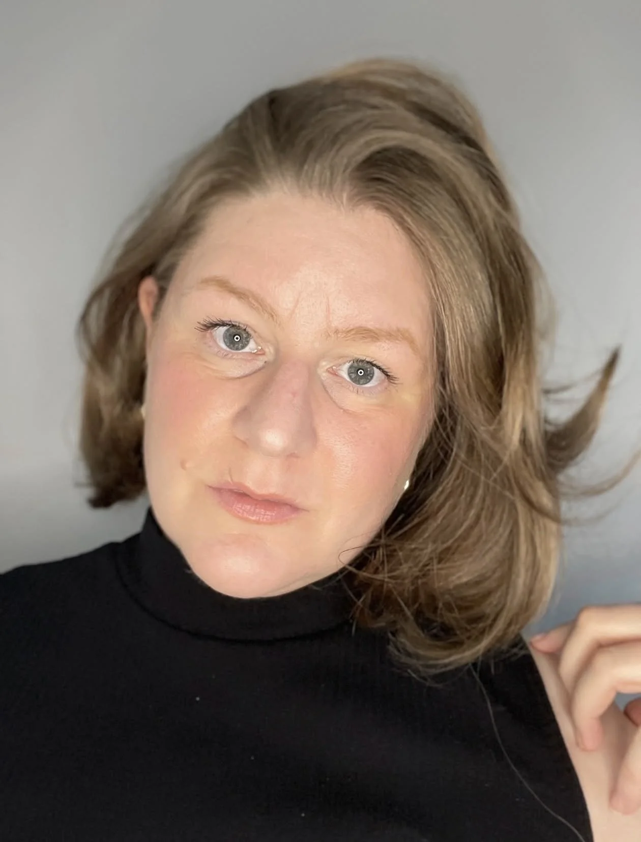 A woman with shoulder-length light brown hair, blue eyes, and fair skin wearing a black turtleneck, looking at the camera with a neutral expression.