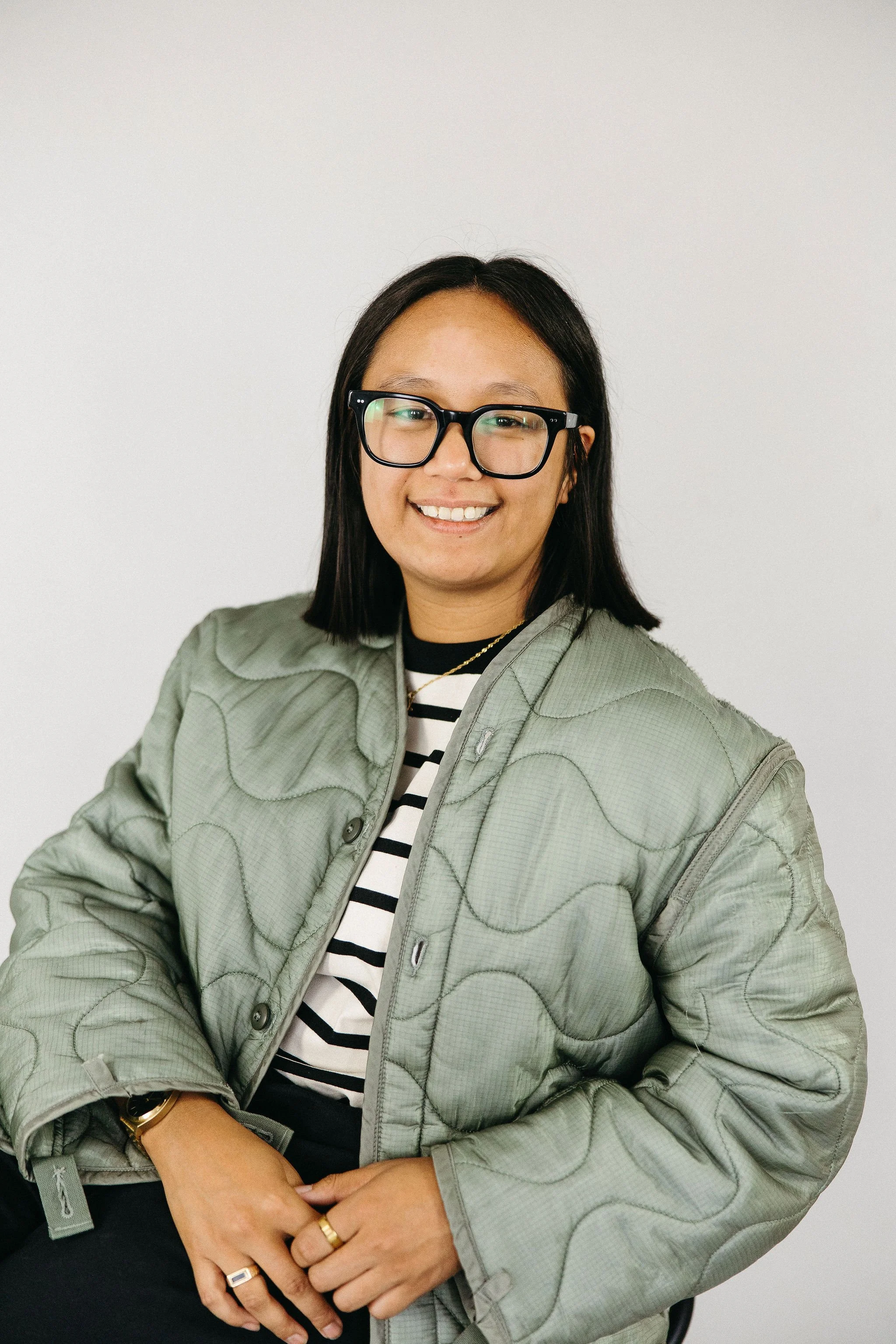 A woman with shoulder length black hair wearing large glasses, a gray quilted jacket, and a striped black and white shirt, sitting against a plain white background.