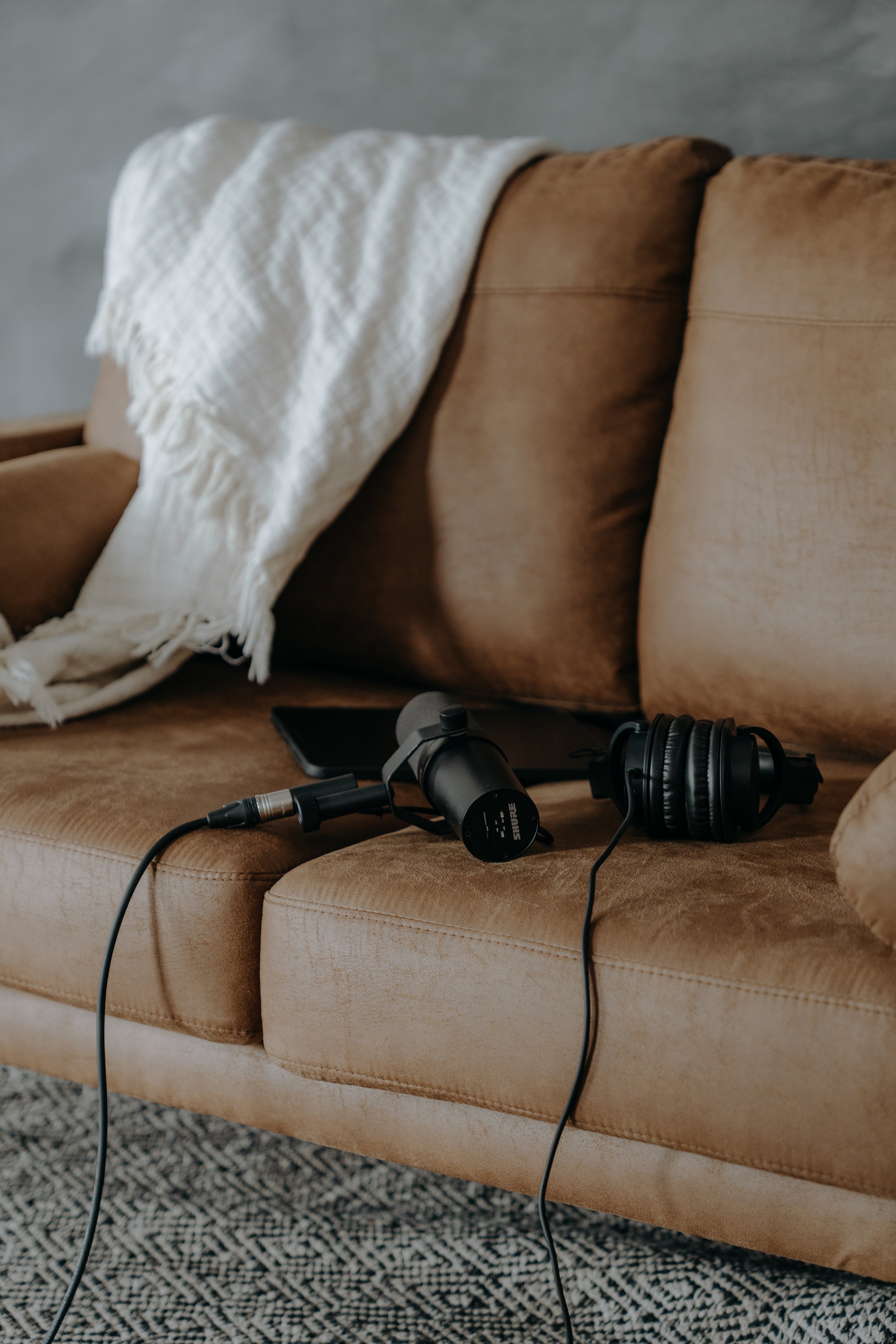 A beige vintage-style sofa with a white throw blanket draped over the backrest. On the sofa's seat, there's a music recording setup with a microphone, a tablet, and a pair of black over-ear headphones.
