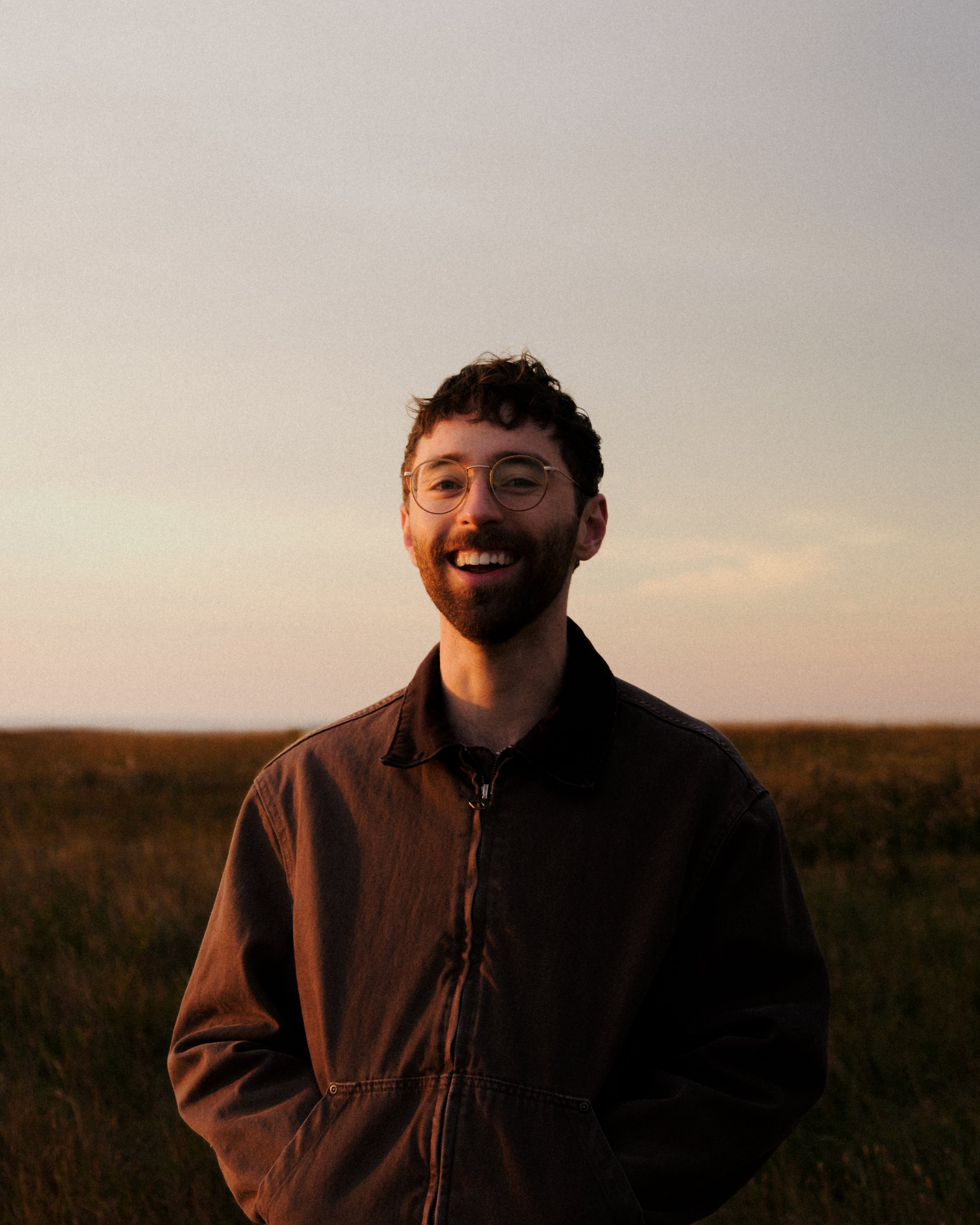 A man with glasses and a beard, smiling, standing outdoors at sunset, wearing a dark jacket in a field.