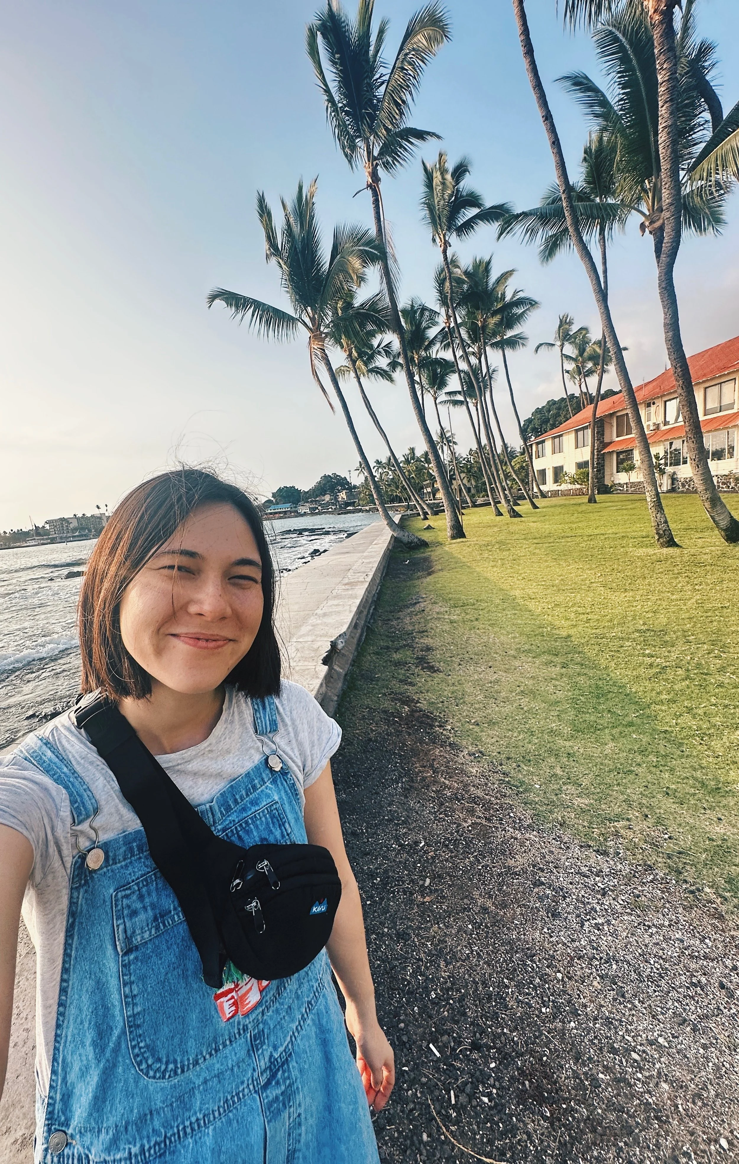 A young woman smiling with closed eyes, taking a selfie near a coastline with palm trees and a grassy area in the background during sunset or late afternoon.