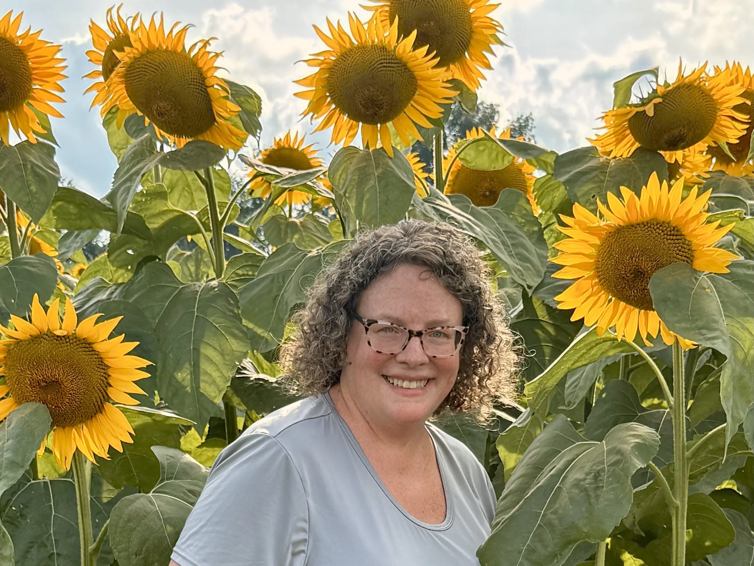 A smiling woman with curly hair and glasses standing among tall sunflower plants with bright yellow petals and large green leaves, under a partly cloudy sky.