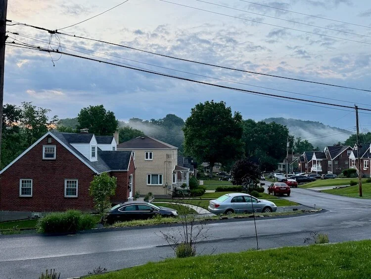 First light of dawn over a Pittsburgh, PA, neighborhood. There is a glimmer of pink-orange sunrise and fog rising above a valley with a creek.