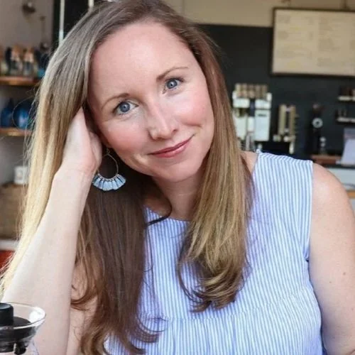 A woman with long blonde hair and blue eyes sitting at a table in a cafe, smiling at the camera.