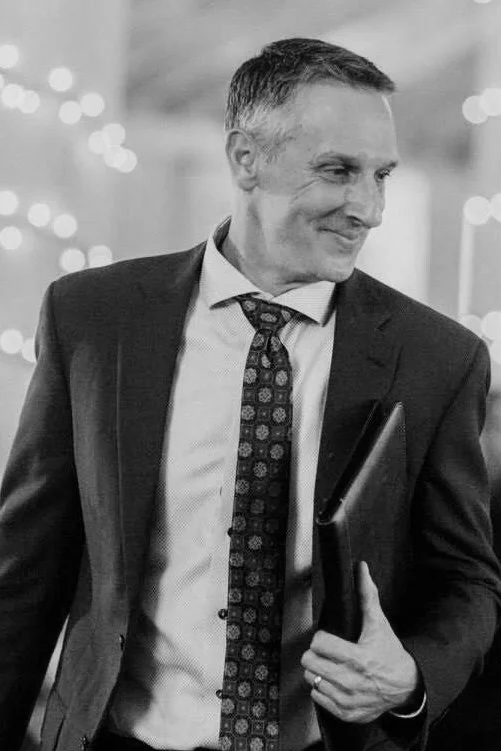 A man in a dark suit and patterned tie holding a black folder, smiling, in a warmly lit indoor setting with blurred decorative lights in the background.