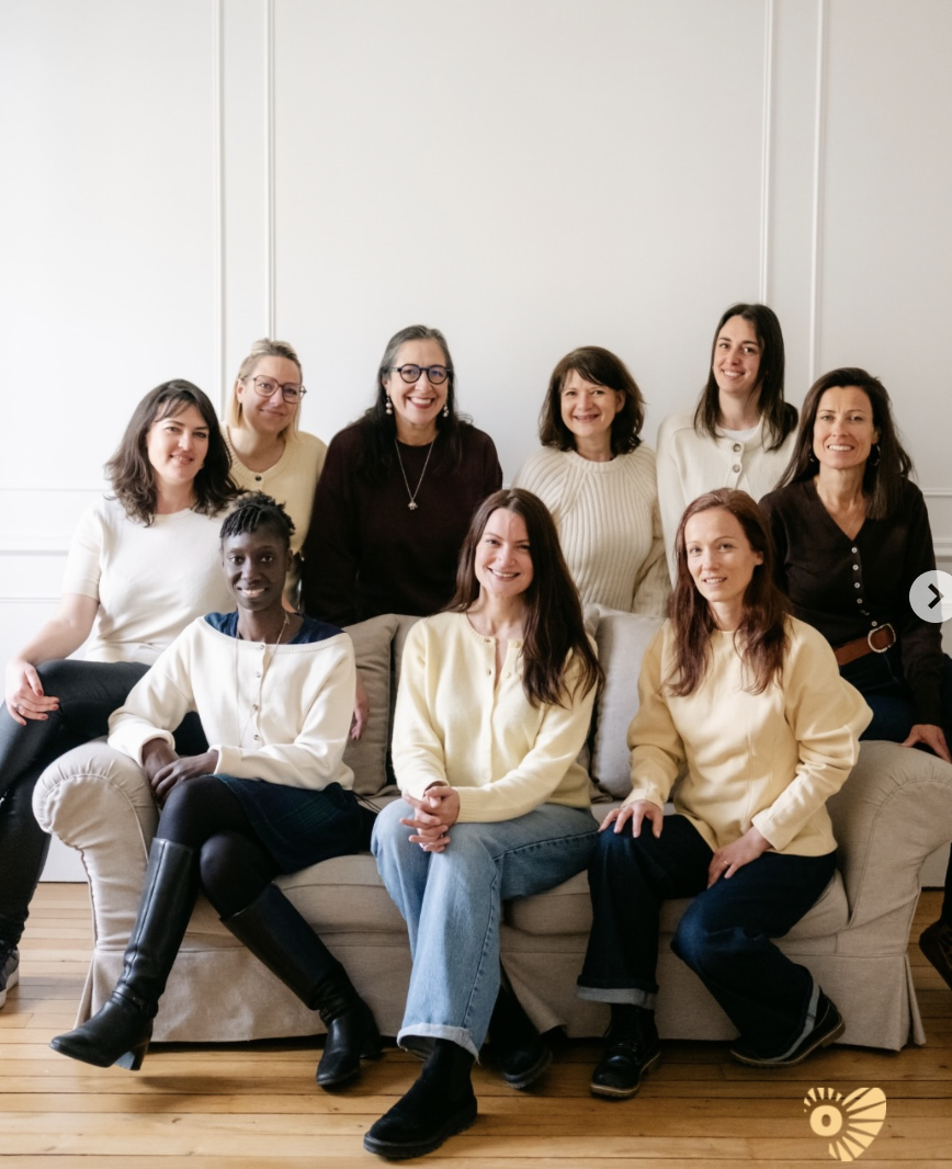 Un groupe de dix femmes souriantes posant dans un salon avec un canapé beige et un sol en bois.