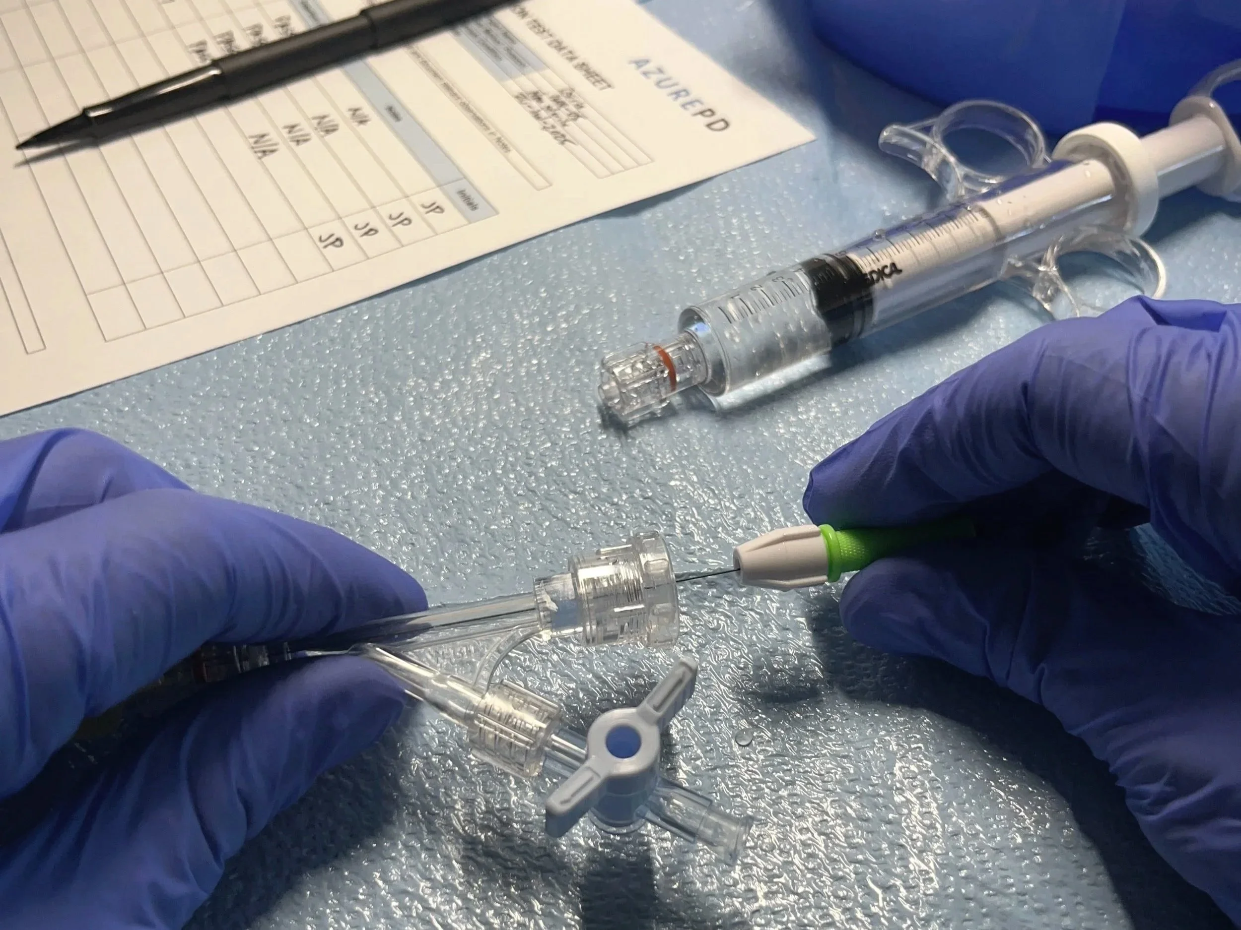 Gloved hands performing an insertion-style test with catheter components, syringe, and datasheet