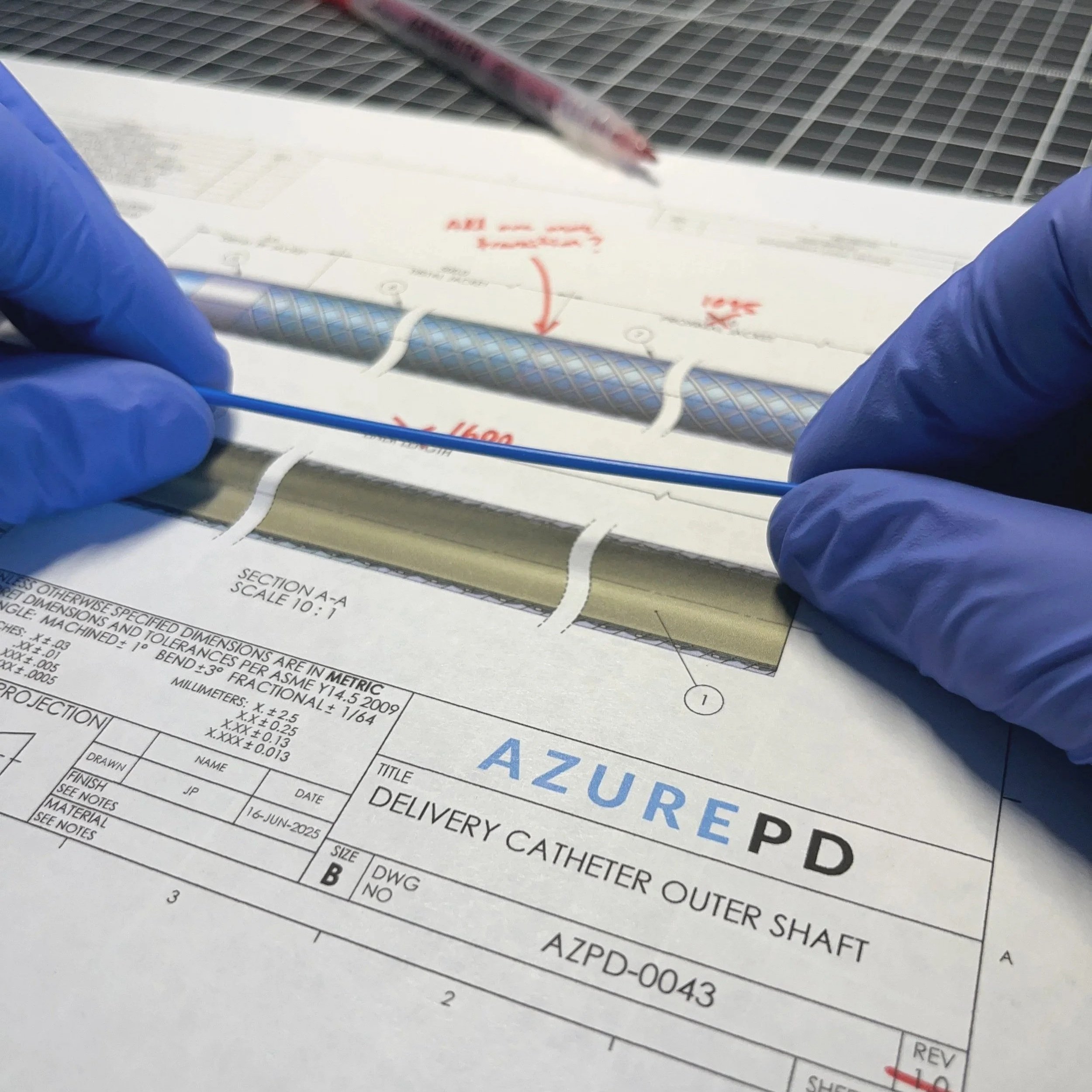 Gloved hands evaluating a catheter shaft prototype over a marked engineering drawing