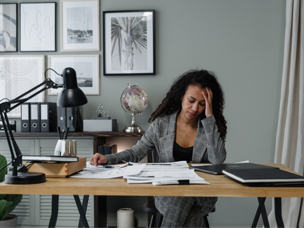 Woman sitting at a desk surrounded by papers, feeling overwhelmed, representing perfectionism stress