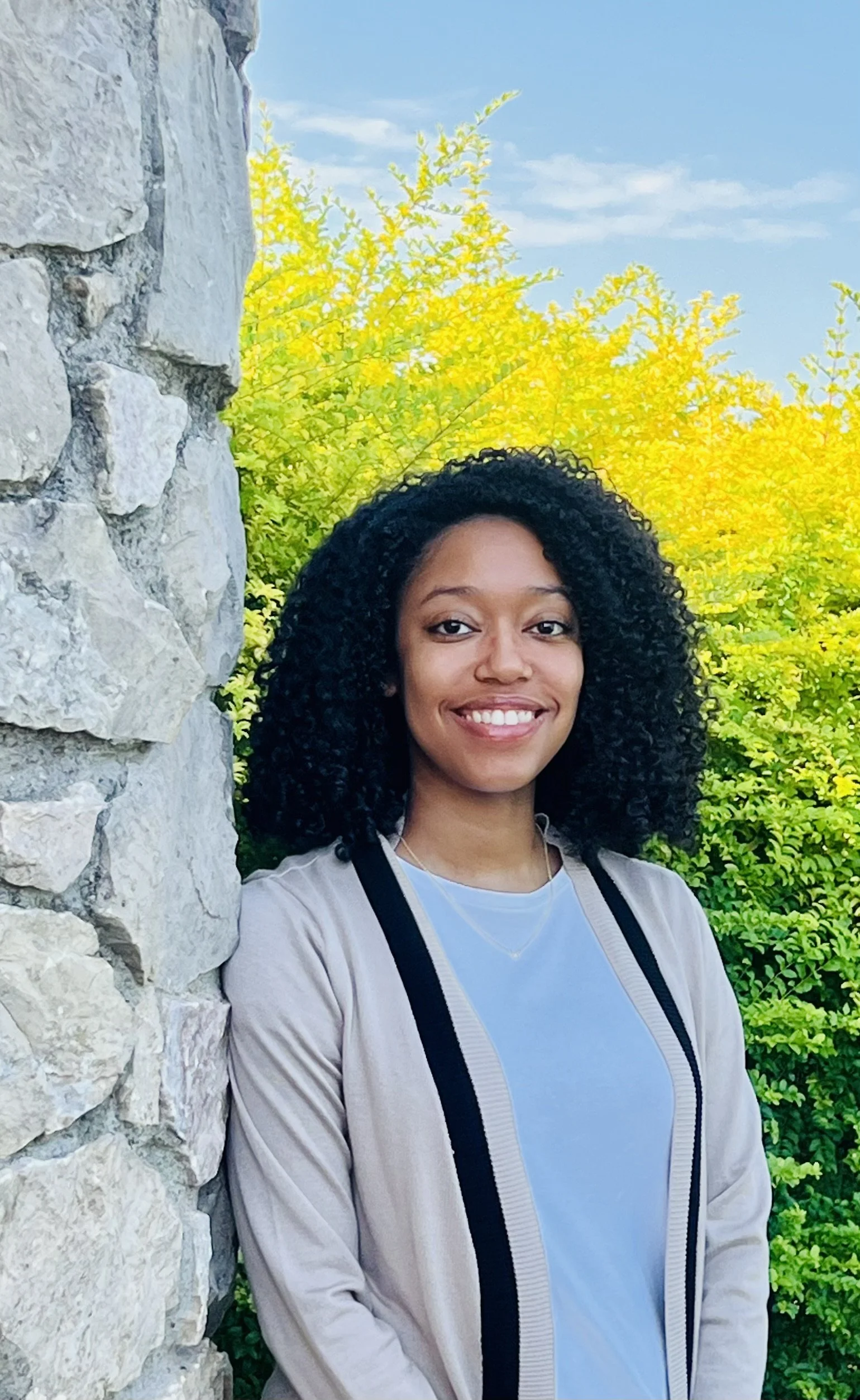A young woman with curly black hair smiling outdoors next to a stone wall with green and yellow trees in the background under a clear blue sky.