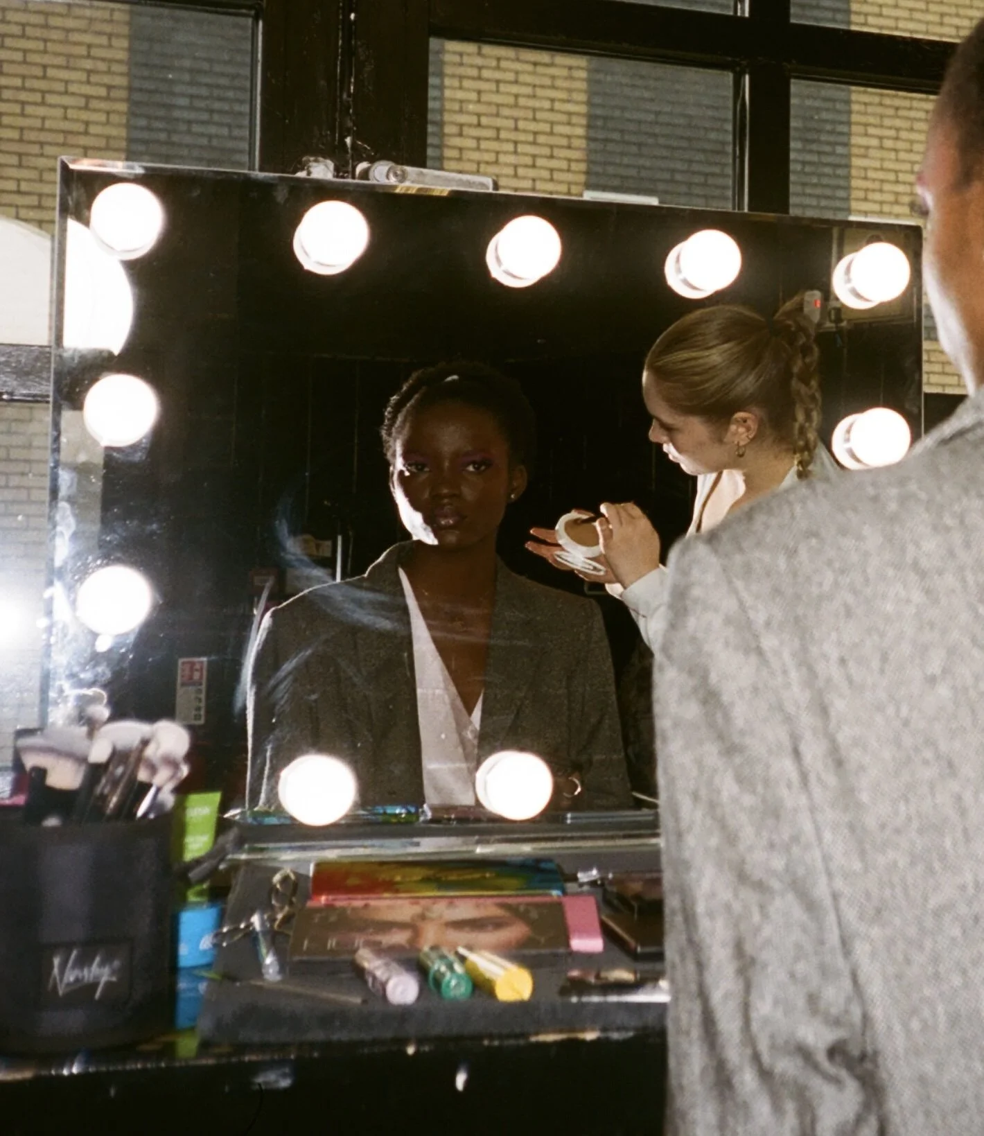 A woman is applying makeup in front of a mirror surrounded by lights. She is focused, and a man with a gray blazer is observing her, with his back to the camera.