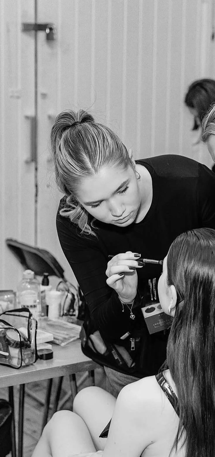 A woman applying makeup to a young girl with long hair in a room with a wooden wall.