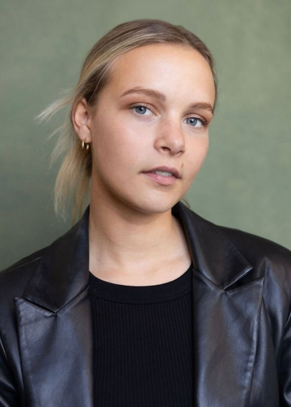 A young woman with light blonde hair, blue eyes, wearing gold hoop earrings, a black top, and a black leather jacket, standing against a green background.