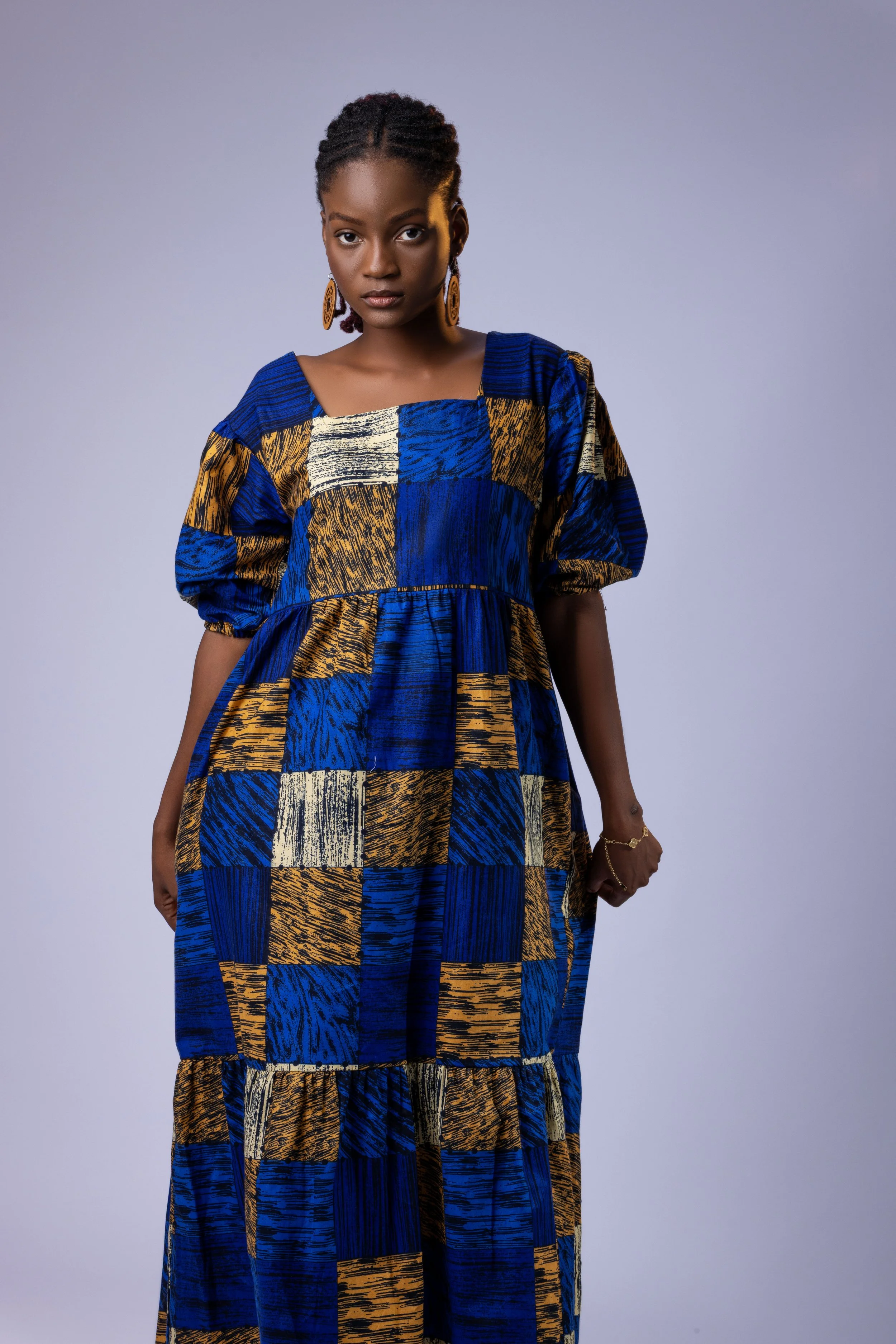 Woman in a colorful, embellished African print dress standing in front of a mirror with a gold frame, posing confidently with her hands on her hips.