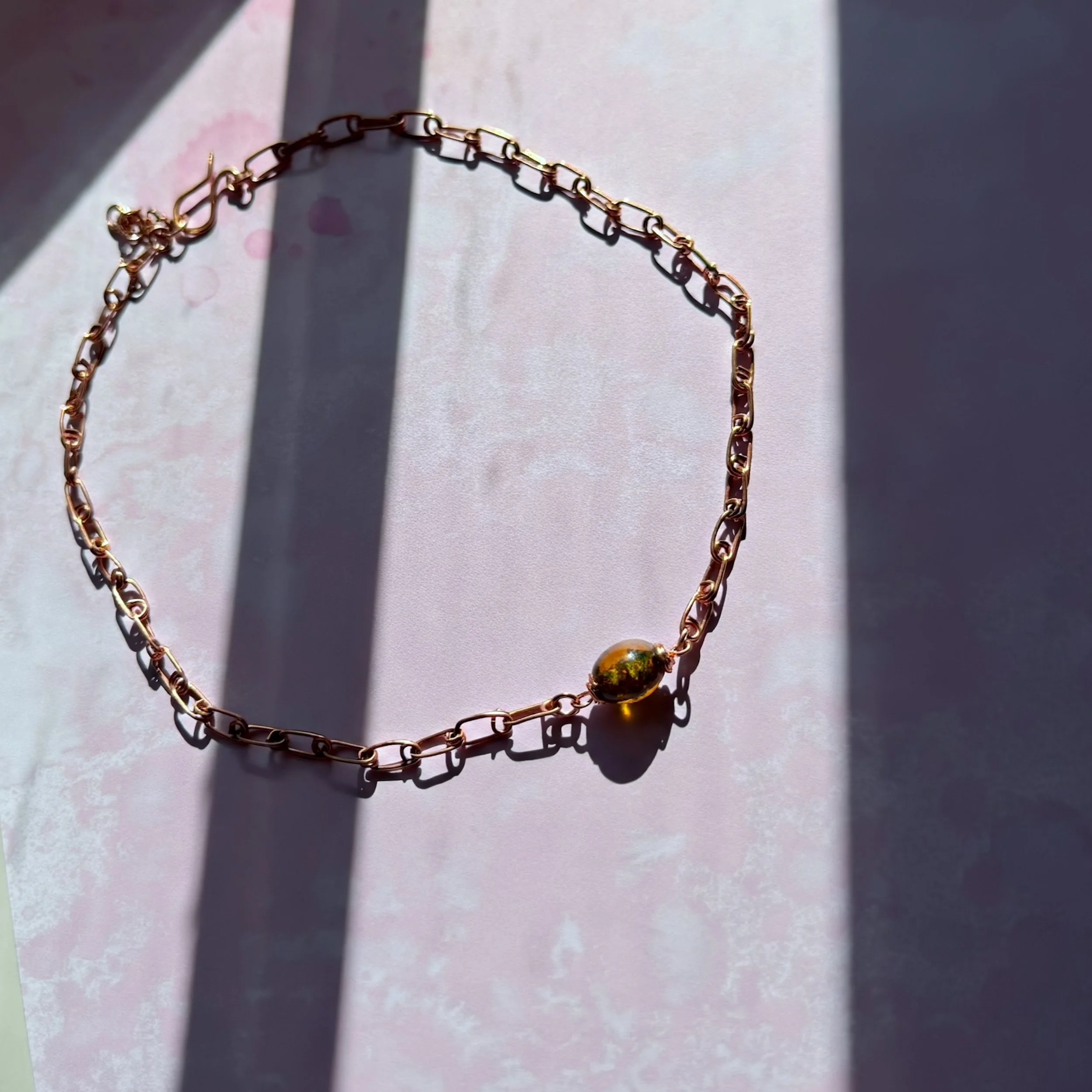 A hand-formed copper safety pin design chain with a sparkly amber lampwork glass bead on a pink background