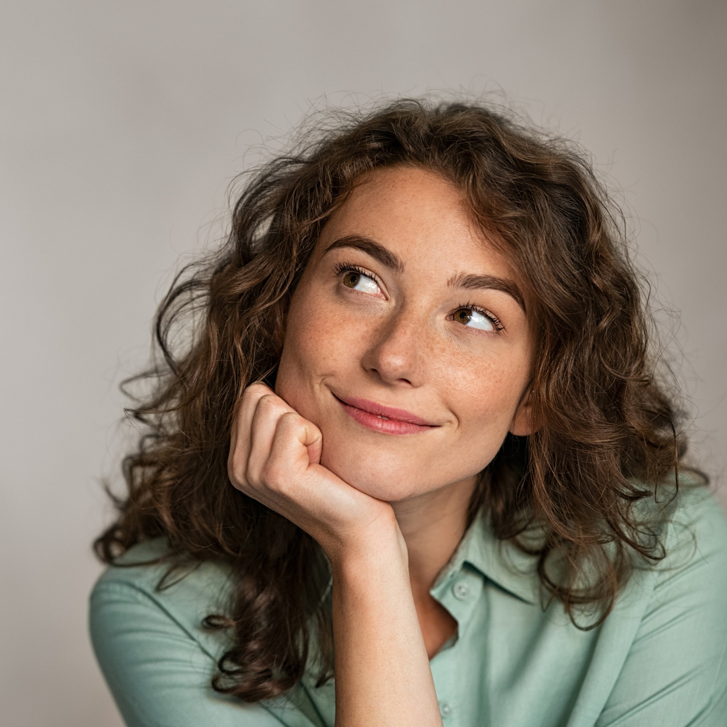 Close-up of a woman with curly brown hair, smiling gently, wearing a green shirt, resting her chin on her hand.