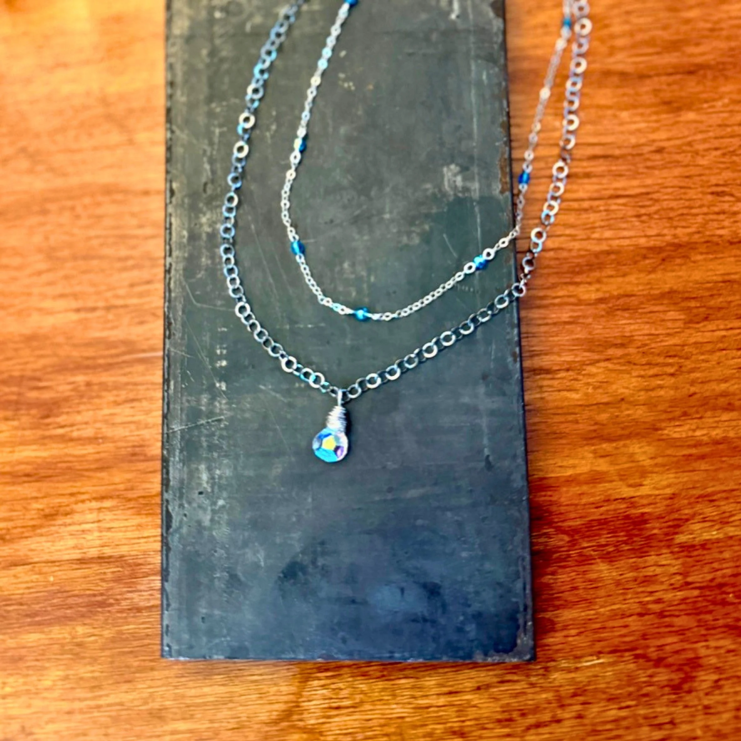 Czech glass Dew Drop Necklace with a silver-finished chain shown with Crystal Wrapped Chain Necklace on metal and wood background