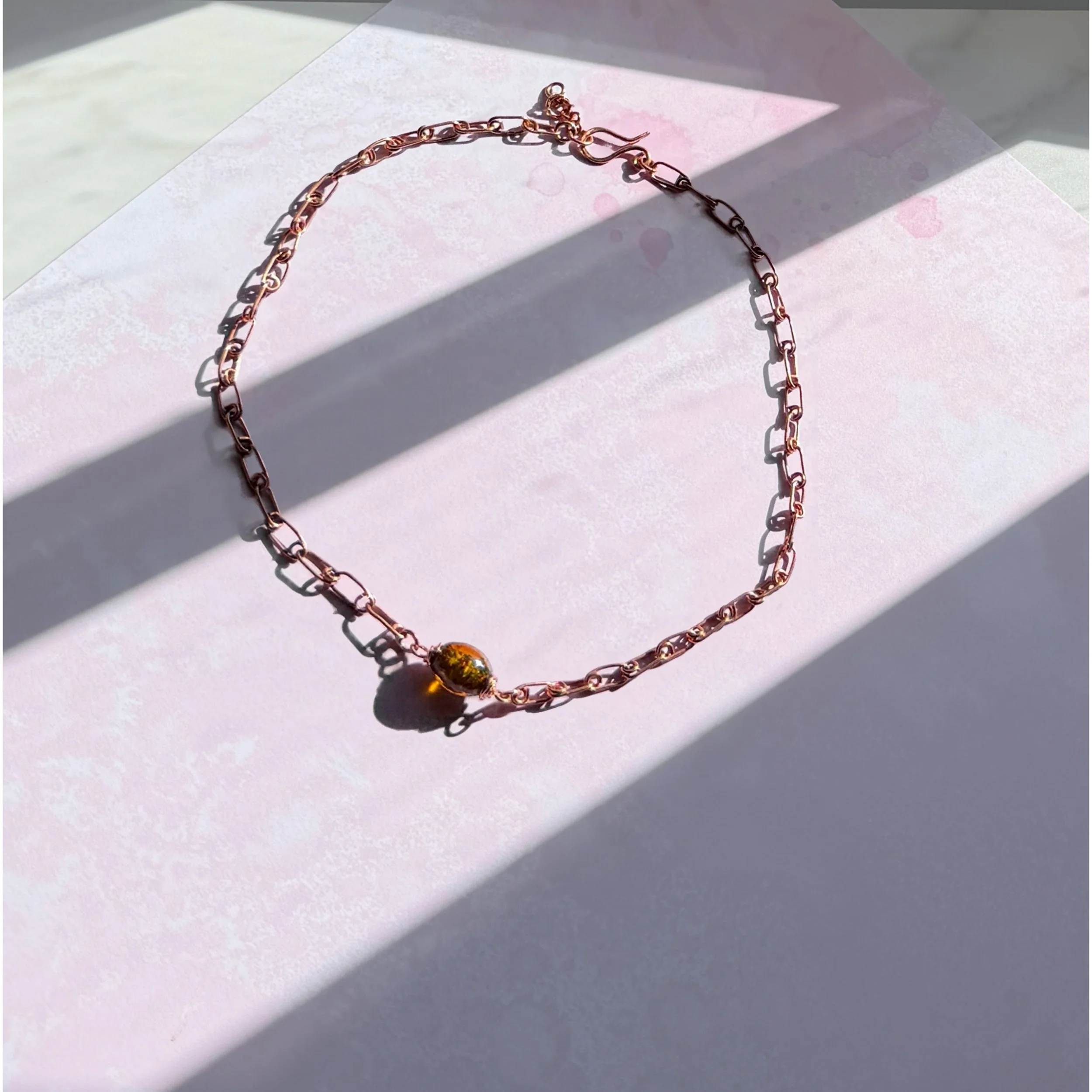A hand-formed copper safety pin design chain with a sparkly amber lampwork glass bead on a pink background