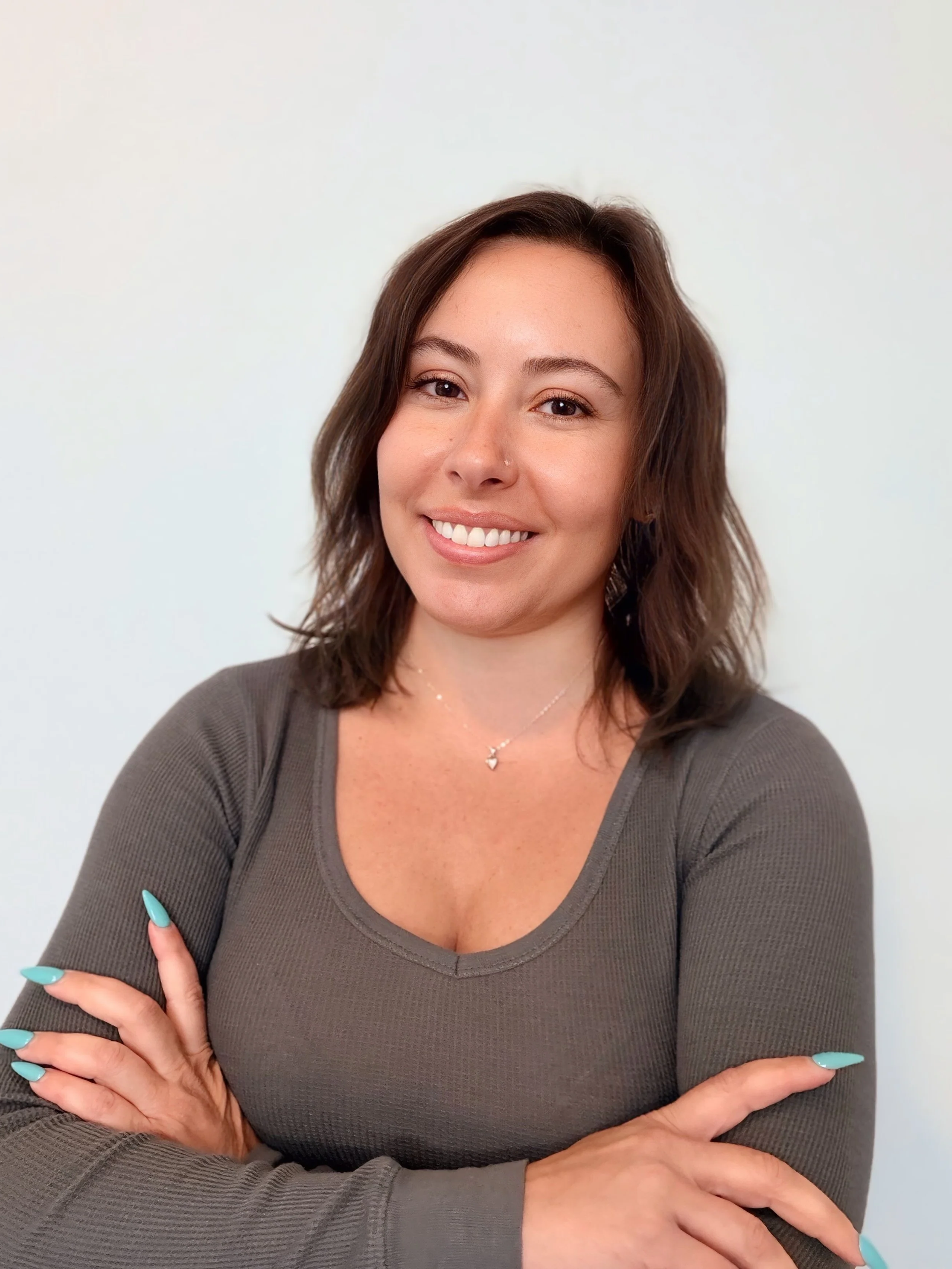 Portrait of a smiling woman with brown hair, wearing a gray top, silver necklace, and turquoise nail polish, standing against a plain white background.