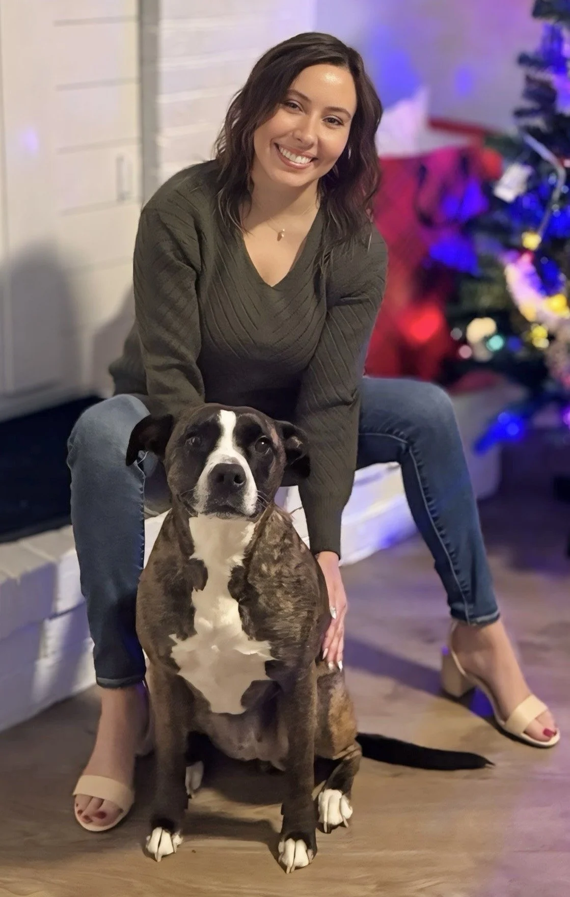 A woman with dark, wavy hair wearing a dark green sweater and jeans is sitting on the floor next to a brindle and white dog. The woman is smiling and holding the dog close, which is sitting and looking at the camera. In the background, there's a decorated Christmas tree with colorful lights, and the setting appears to be indoors, likely during the holiday season.
