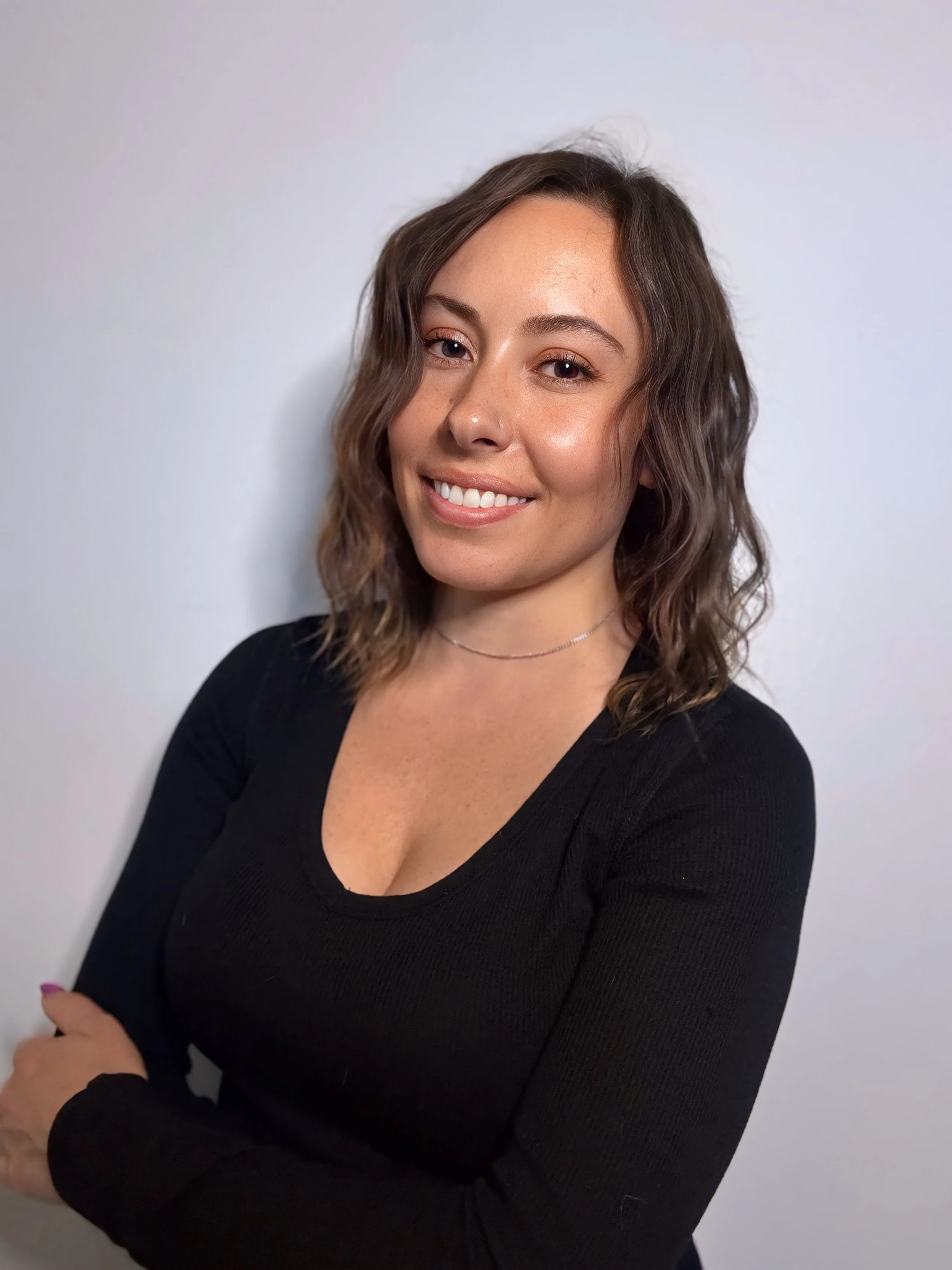 A young woman with shoulder-length wavy brown hair is smiling, wearing a black V-neck top and a delicate necklace, standing against a plain light gray background.