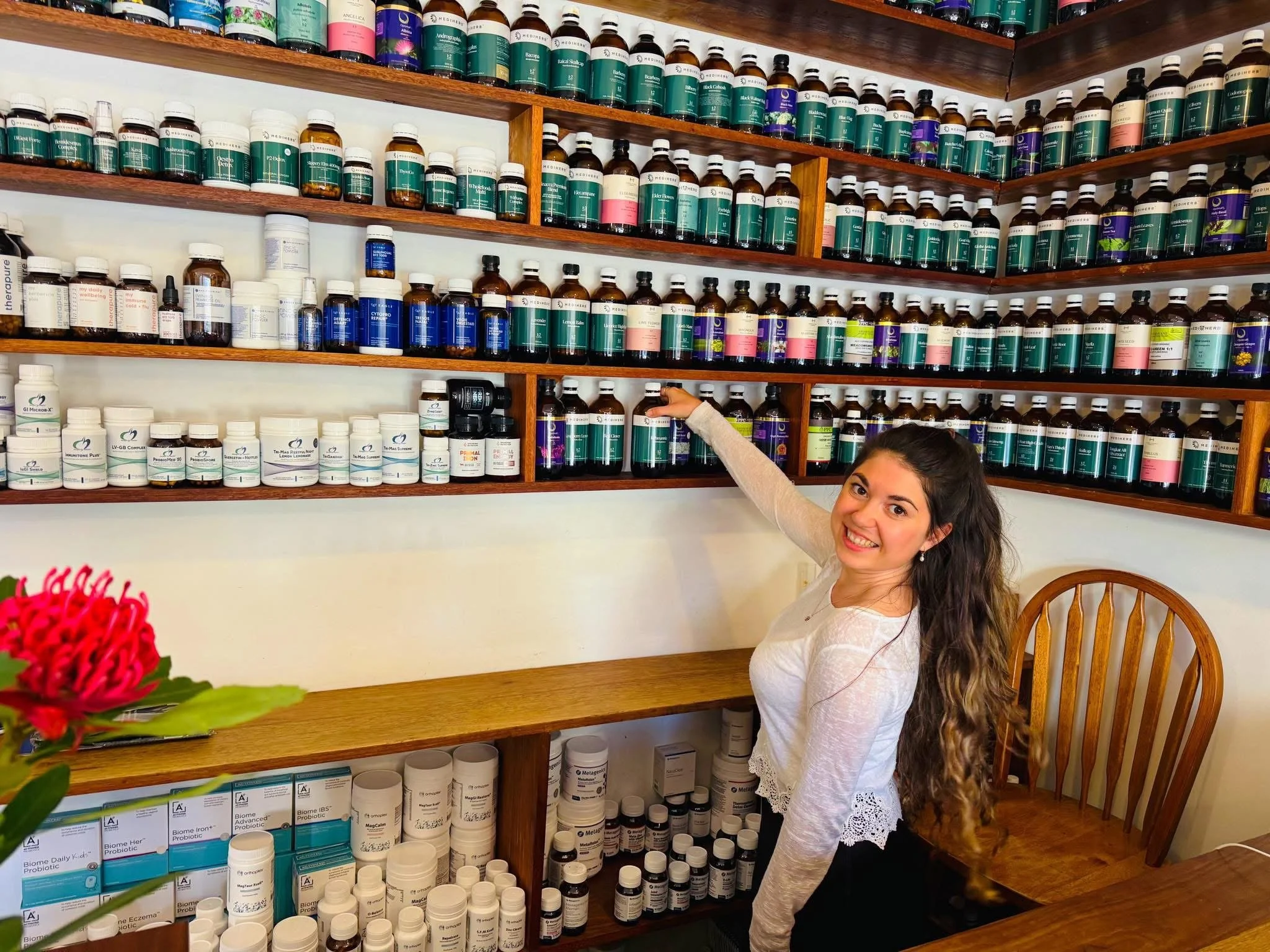 A young woman with long, curly hair smiling and sitting at a wooden desk in front of shelves filled with various bottles of medicine or supplements in a pharmacy or health store.