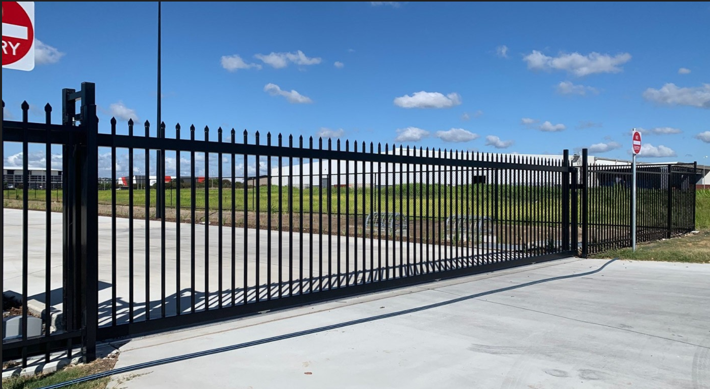 Black metal fence with gate, no entry signs, concrete pavement, grassy area, and clear blue sky.