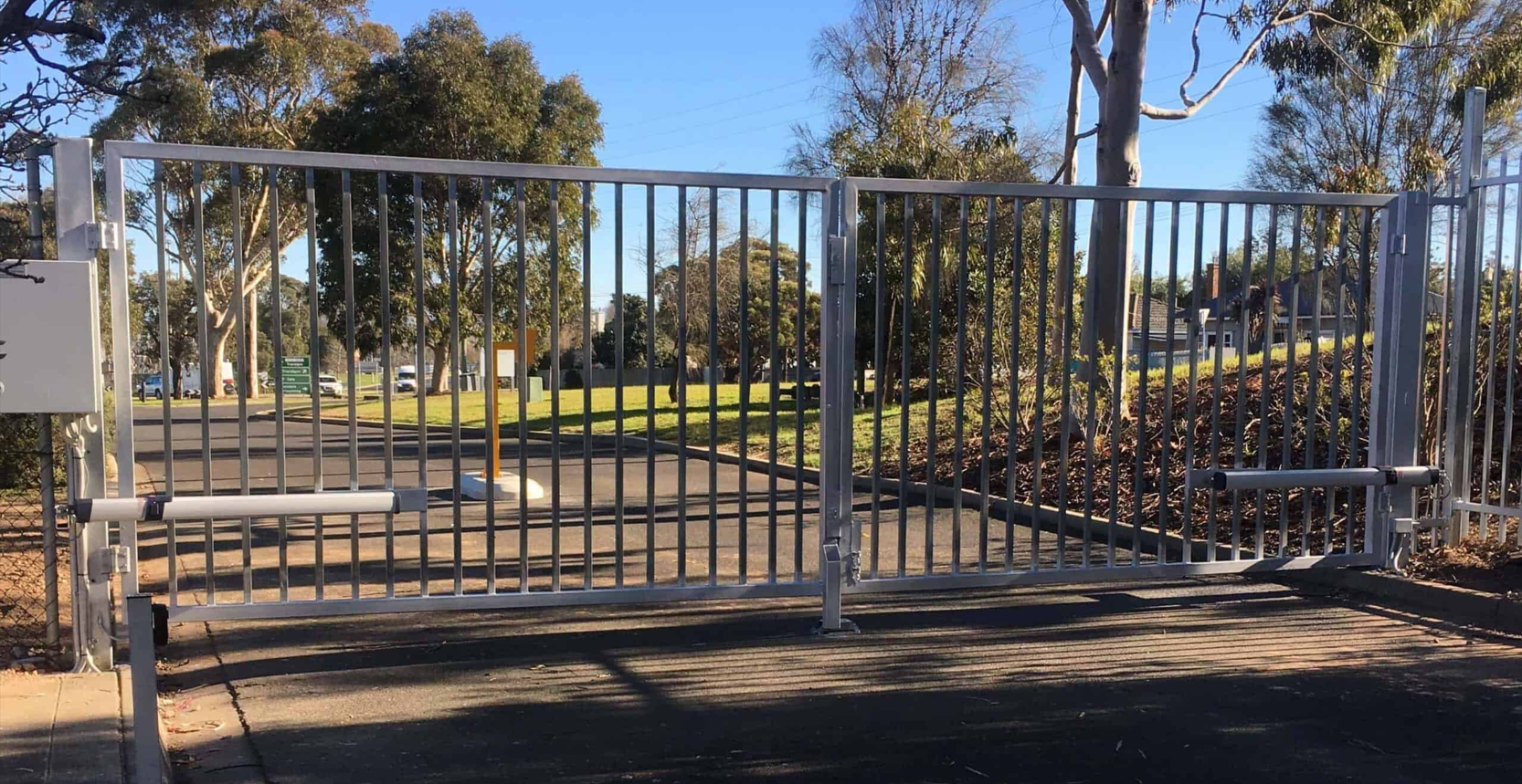 Metal security gate closed on a paved driveway surrounded by trees and grass.