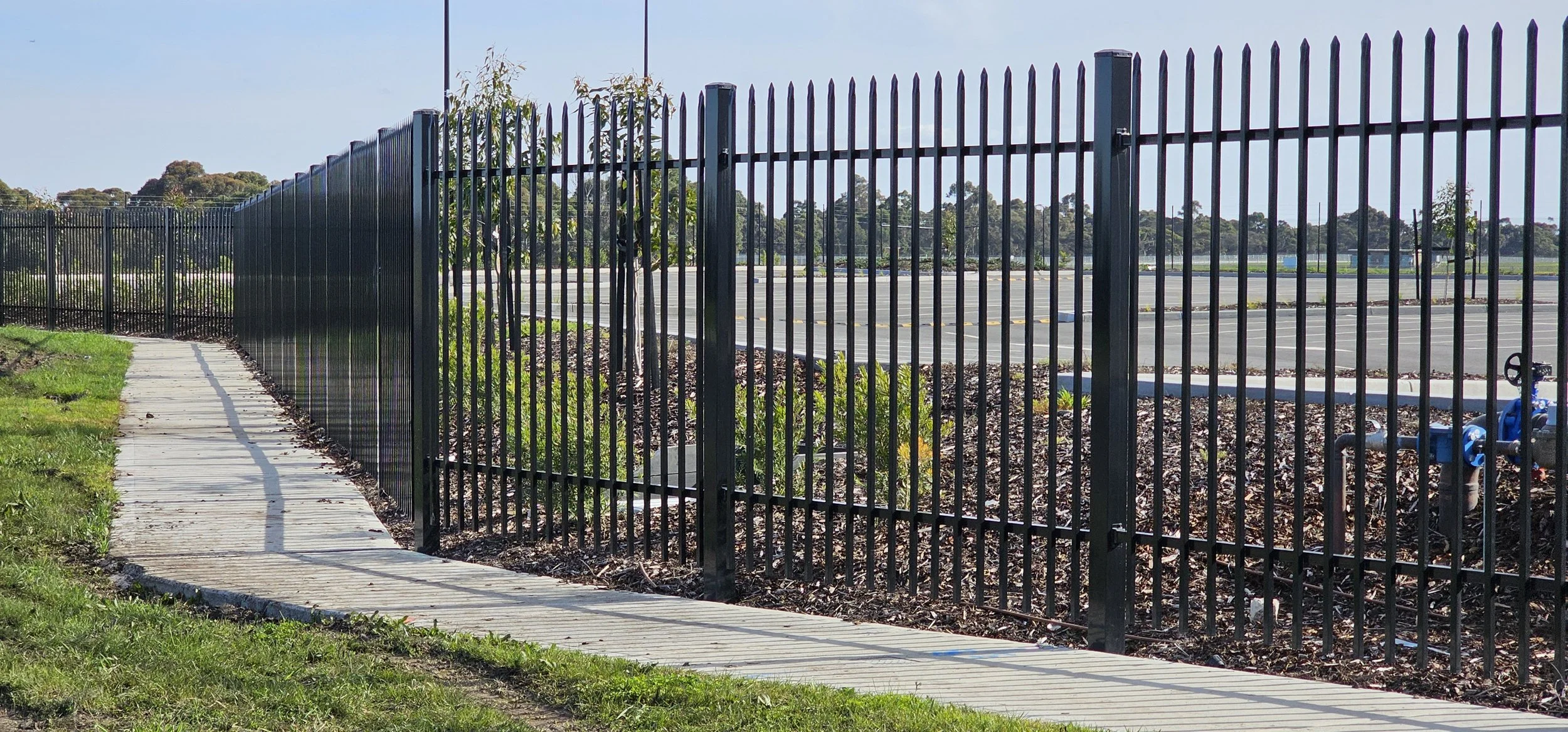 Black metal fence along concrete path with grass, shrubs, and a parking lot in the background.