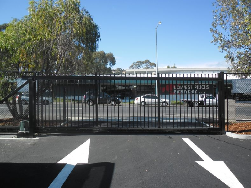 Sliding black metal gate in a parking lot, with a street and cars in the background.