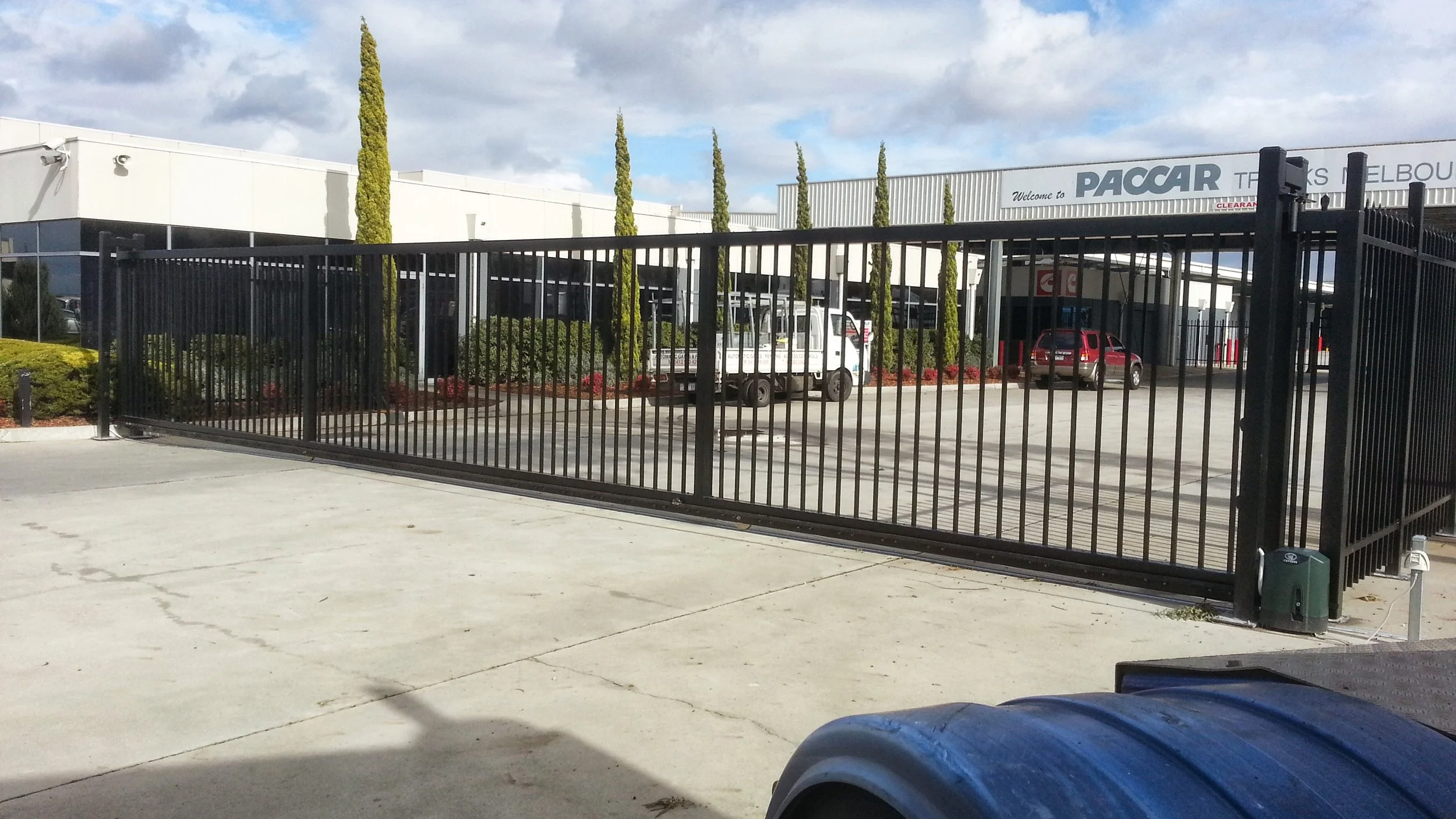 Large sliding black metal gate in front of an industrial building with trucks and cars visible inside. The building is marked 'PACC.' Tall trees and shrubs line the property. Sky is partly cloudy.