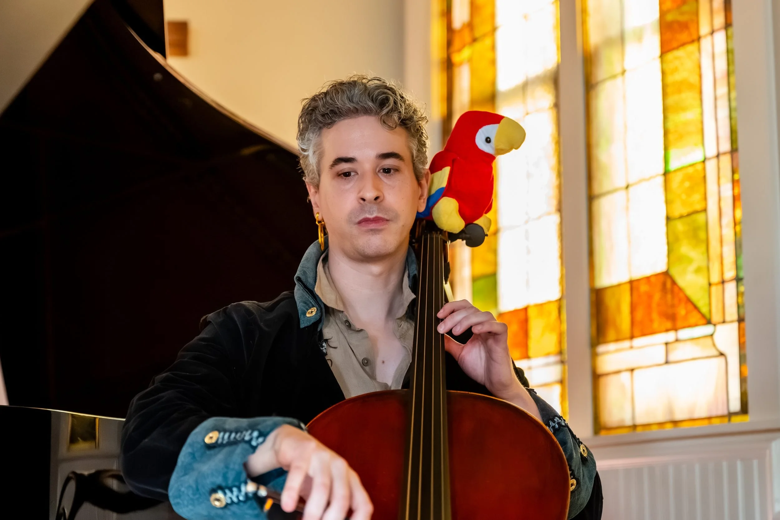 Cellist in a pirate coat with a red toy parrot perched on his cello, stained glass behind him at the Frisco Heritage Center Chapel