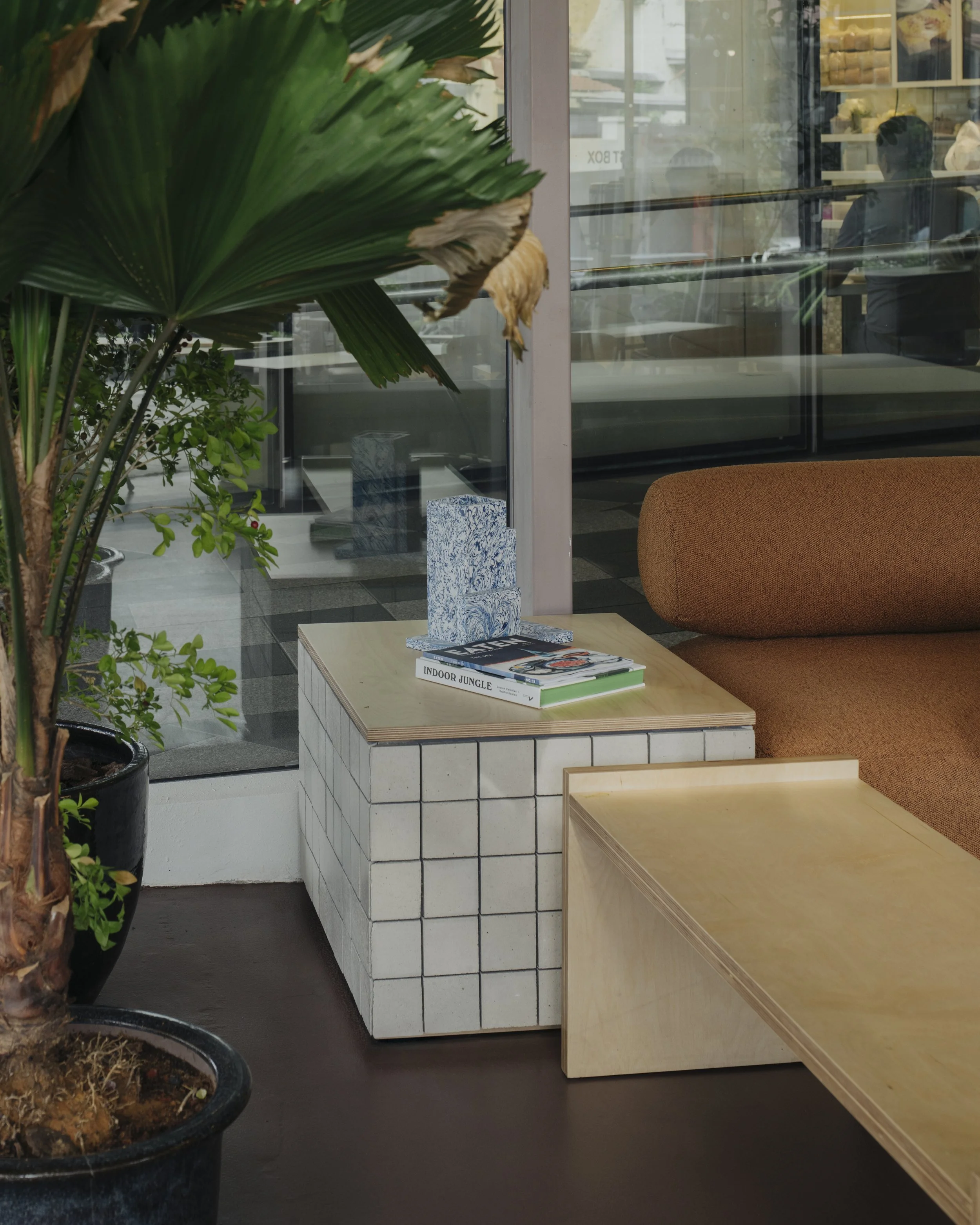 Interior of a cafe or lounge with potted plants, a wooden table, a brick and wood side table with magazines, and a brown cushioned couch, with a glass wall showing the street outside.