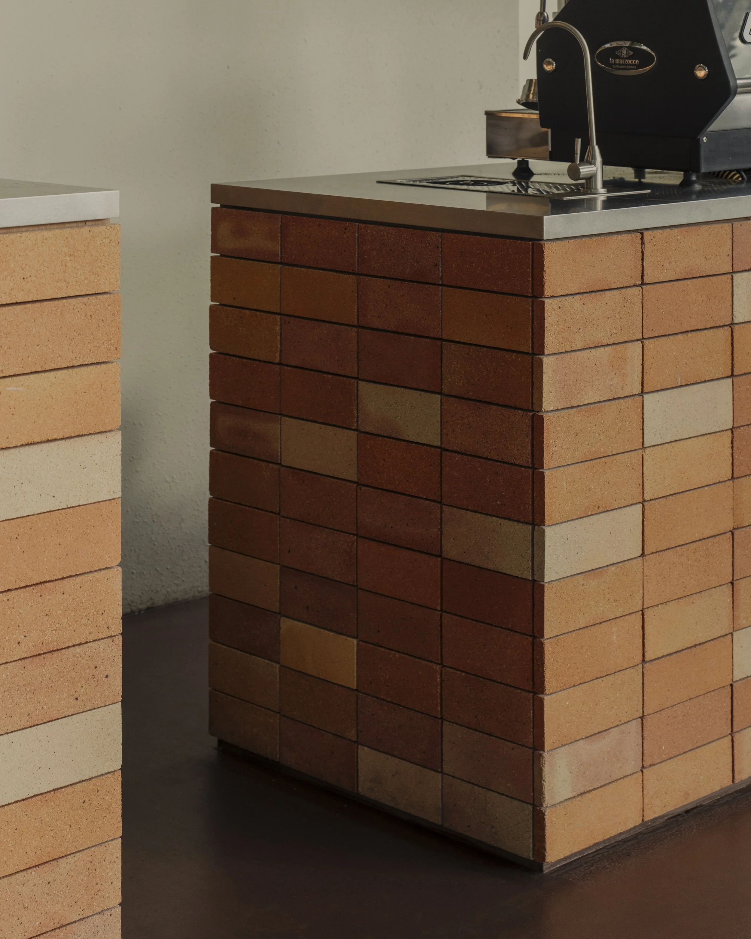 Close-up of a brick kitchen counter with a stainless steel countertop, a black espresso machine, and a sink with a faucet.