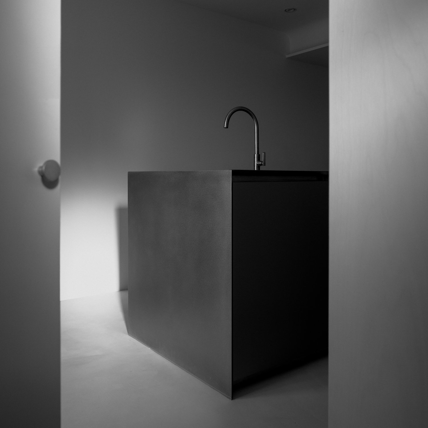 Black minimalist kitchen island with a sink and a curved faucet, viewed through an open door in black and white.
