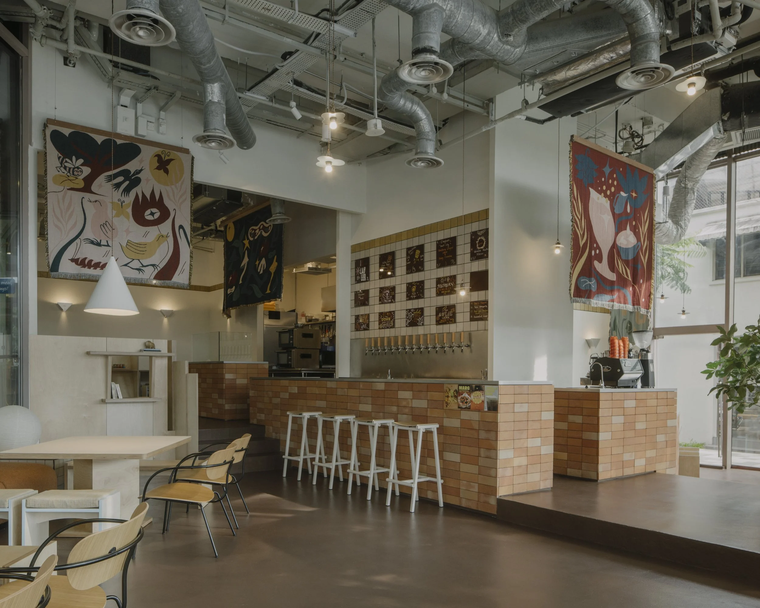 Modern cafe interior with brick counter, white bar stools, and colorful wall tapestries, large windows with plants, and ceiling with exposed ductwork.