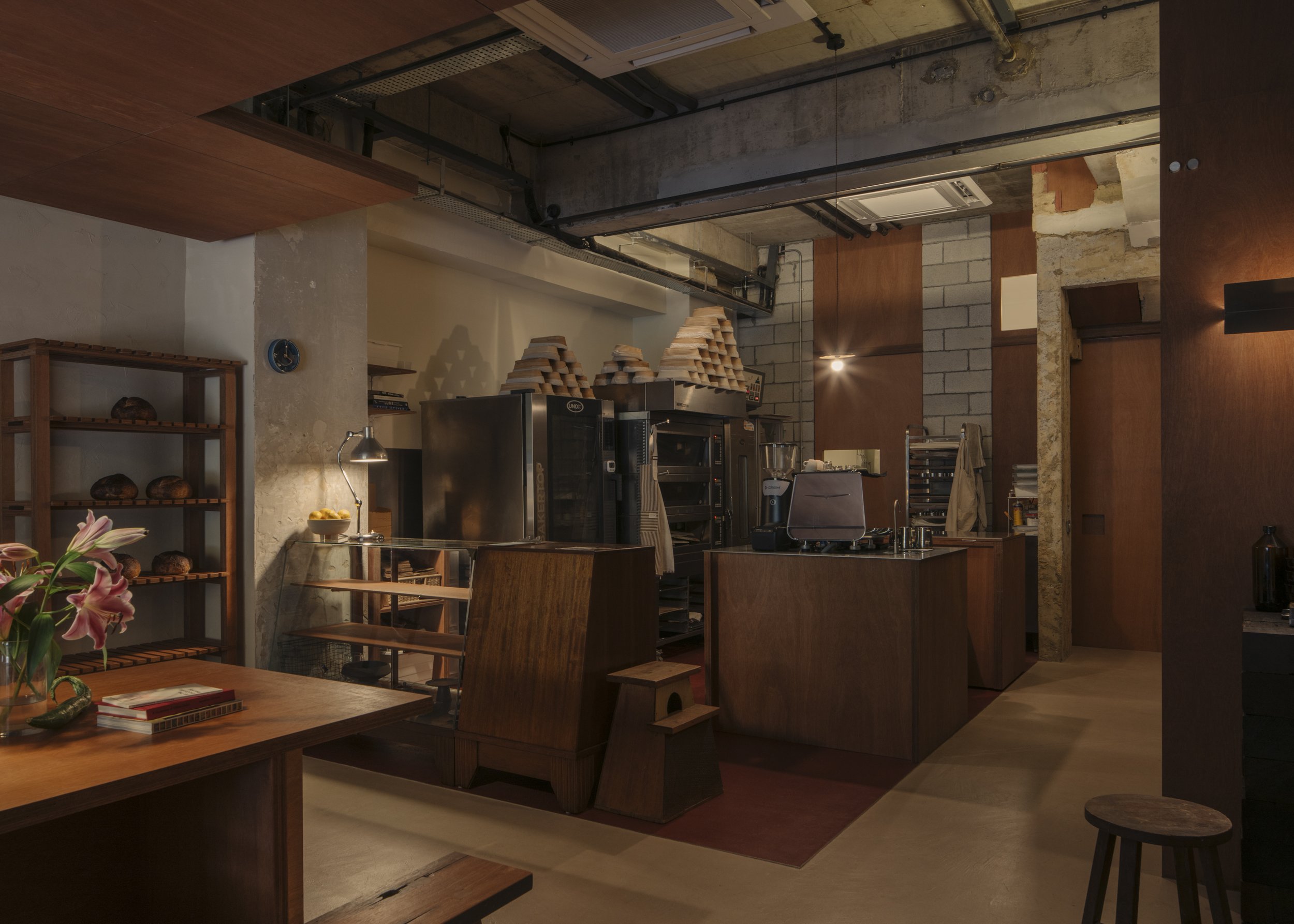 Interior of a coffee shop or bakery with wooden furniture, shelves of baked goods, a coffee counter, and industrial-style decor.