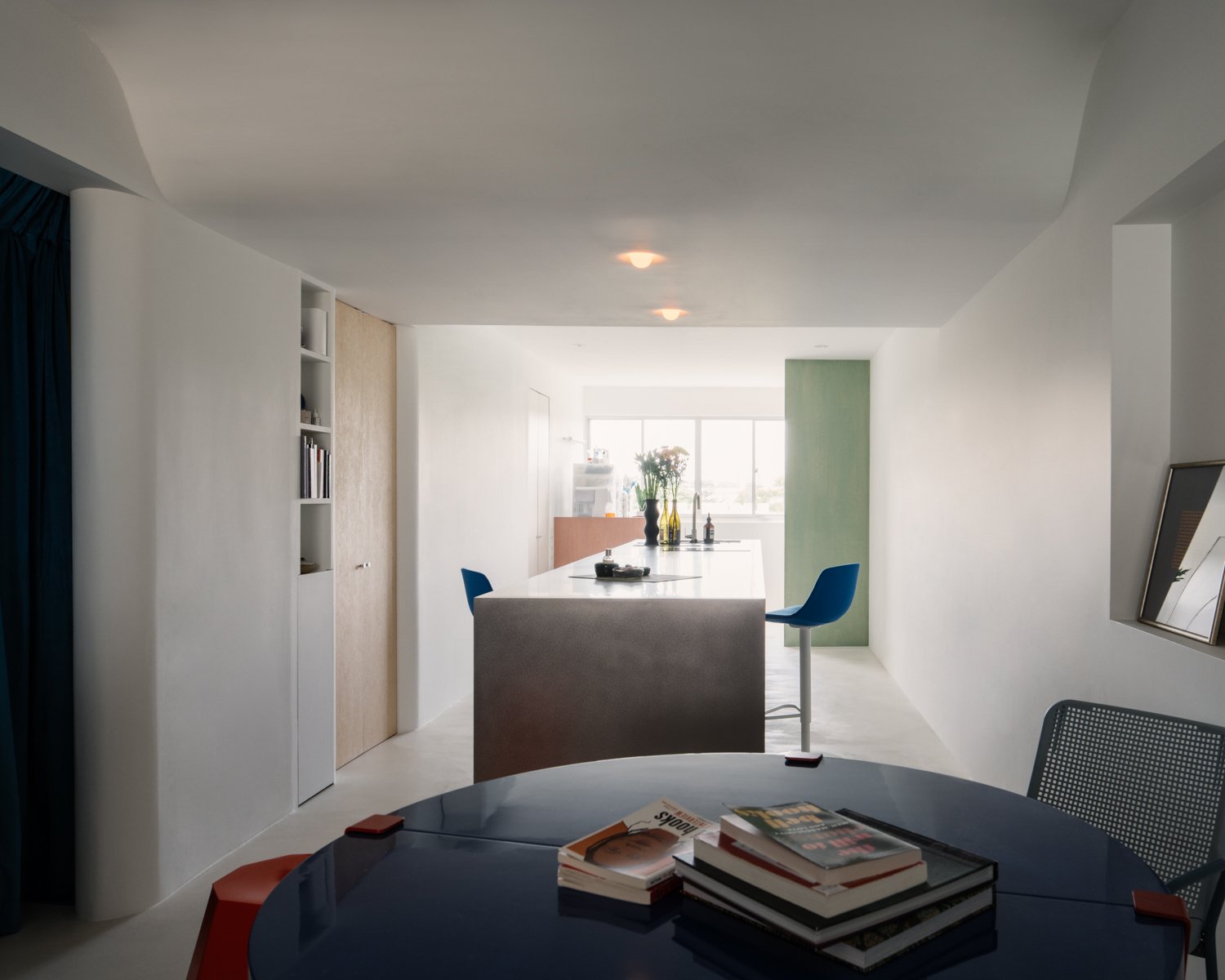 Modern open-plan kitchen and dining area with a round black table, books, and a bright background with a bar counter and blue bar chairs near large windows.