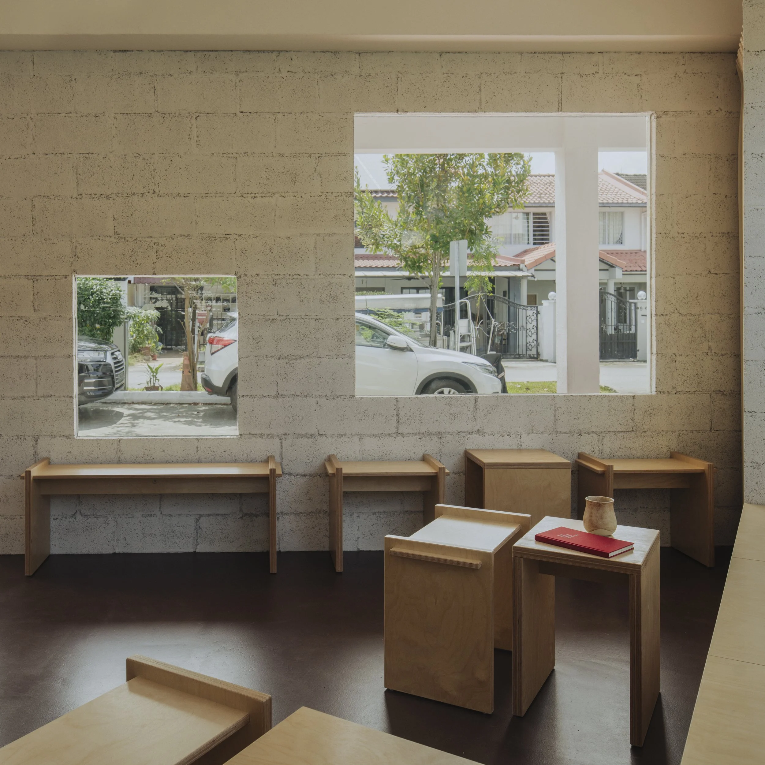 Interior of a room with a concrete block wall with three openings showing outside view of parked cars, trees, and residential houses; minimalistic wooden furniture including benches and tables, with a red book and a small ceramic vase on one table.
