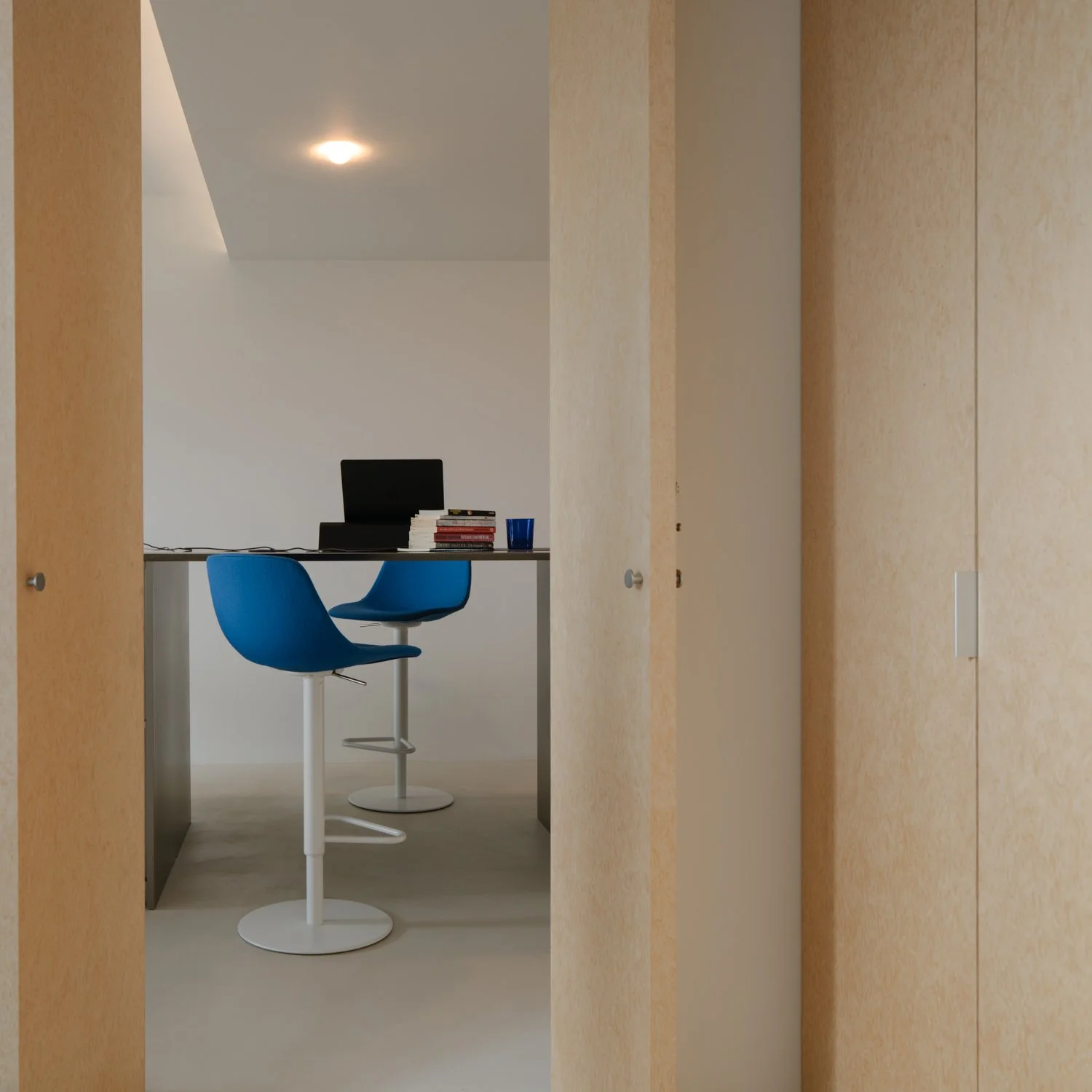 Interior view through a doorway showing a small office or study area with a white wall, a desk with a computer, stack of books, and a blue cup, along with two blue chairs.