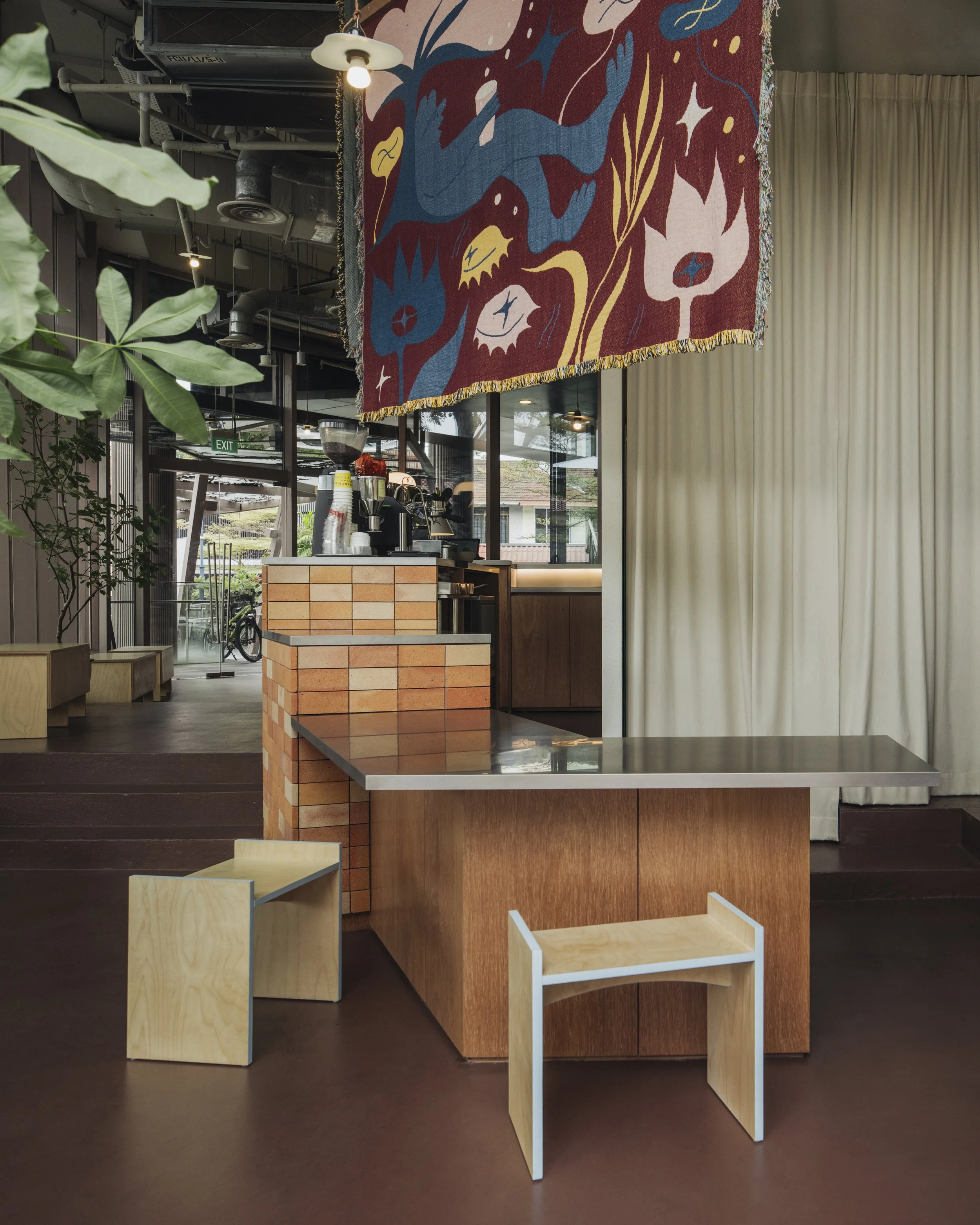 Interior of a cafe with a dark brown floor, wooden furniture, and a small counter with brickwork. There are two small wooden chairs, one in natural wood and the other painted white. A colorful textile art piece with abstract shapes hangs from the ceiling above the counter. Large plants, glass windows, and curtains are visible in the background.