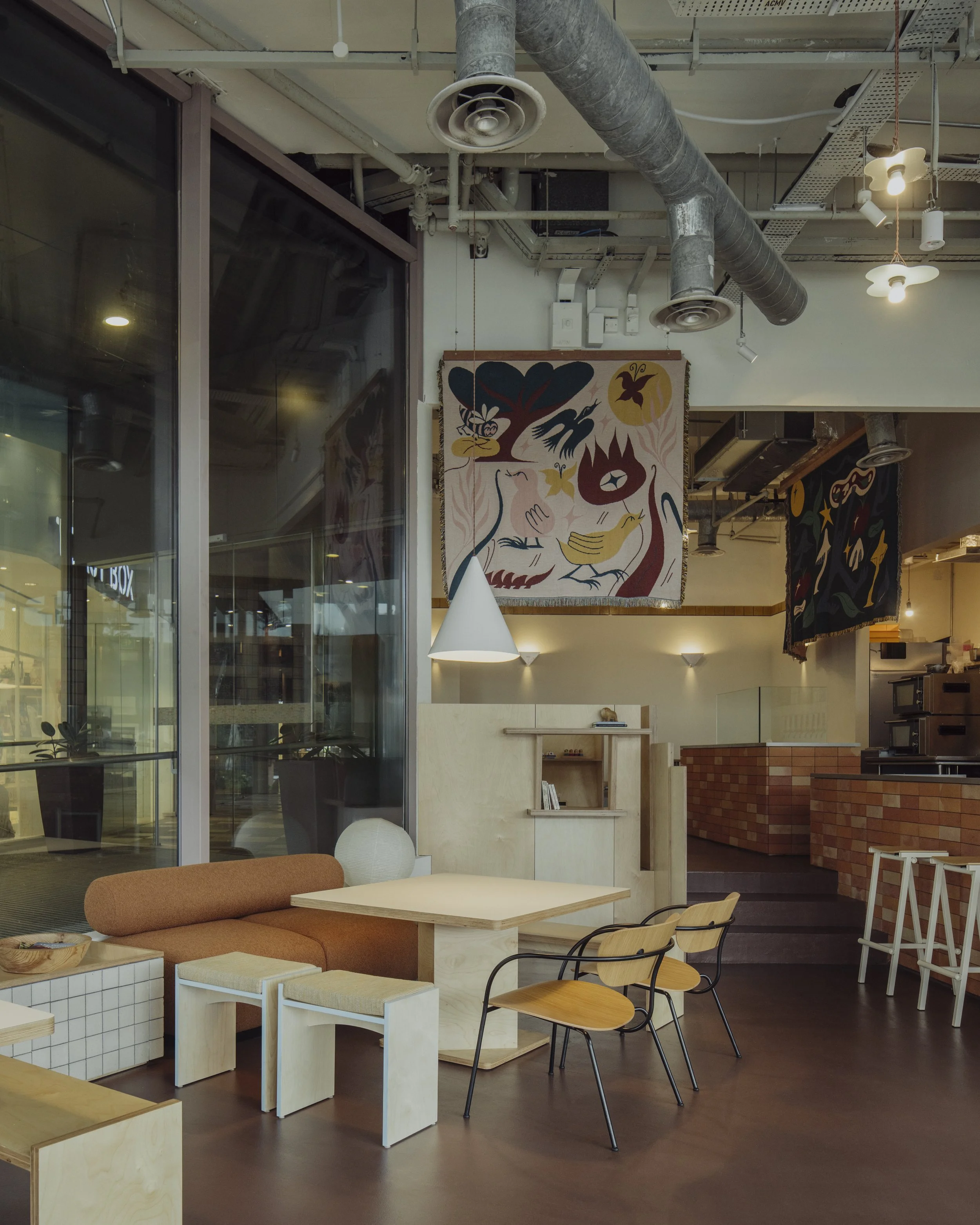 Interior of a modern cafe with a cozy seating area, light-colored wooden furniture, orange cushioned bench, black and wooden chairs, decorative hanging textiles, and industrial ceiling with exposed ducts and lighting.