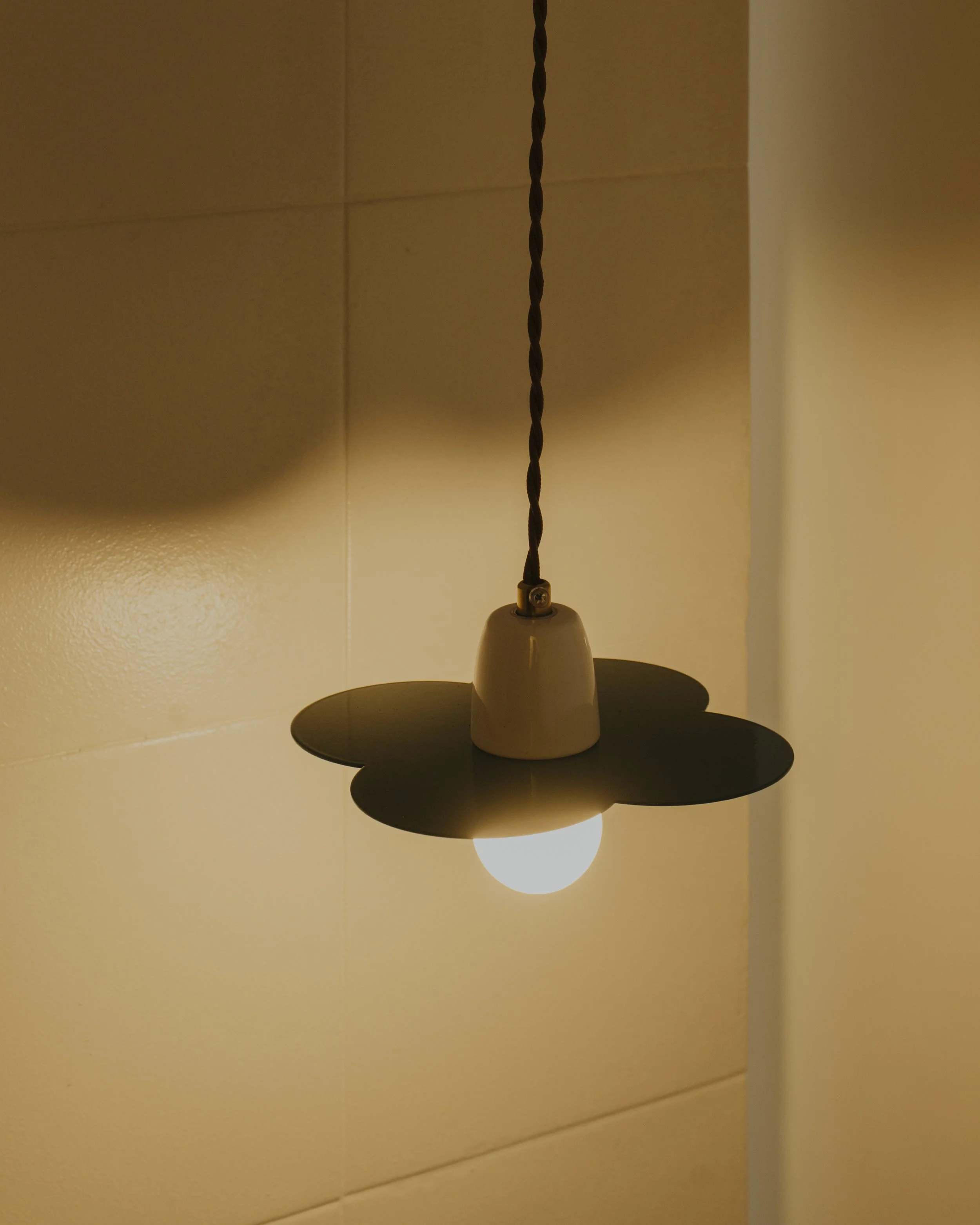 A ceiling light fixture with a black flower-shaped shade and a white ceramic base, hanging from a twisted black cord on a beige tiled wall.