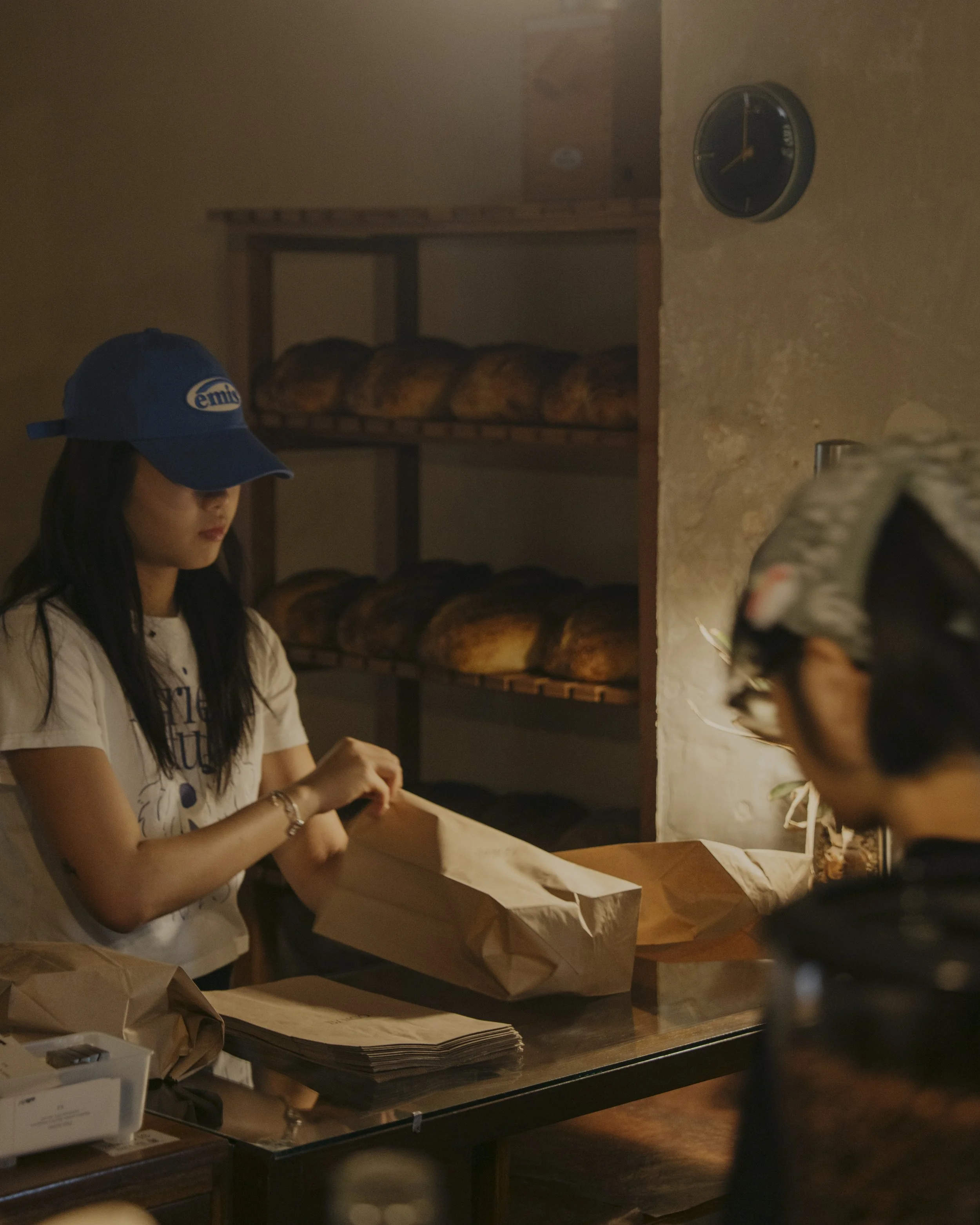 A woman wearing a blue cap and white shirt is packing paper bags at a bakery counter, with shelves of bread in the background.